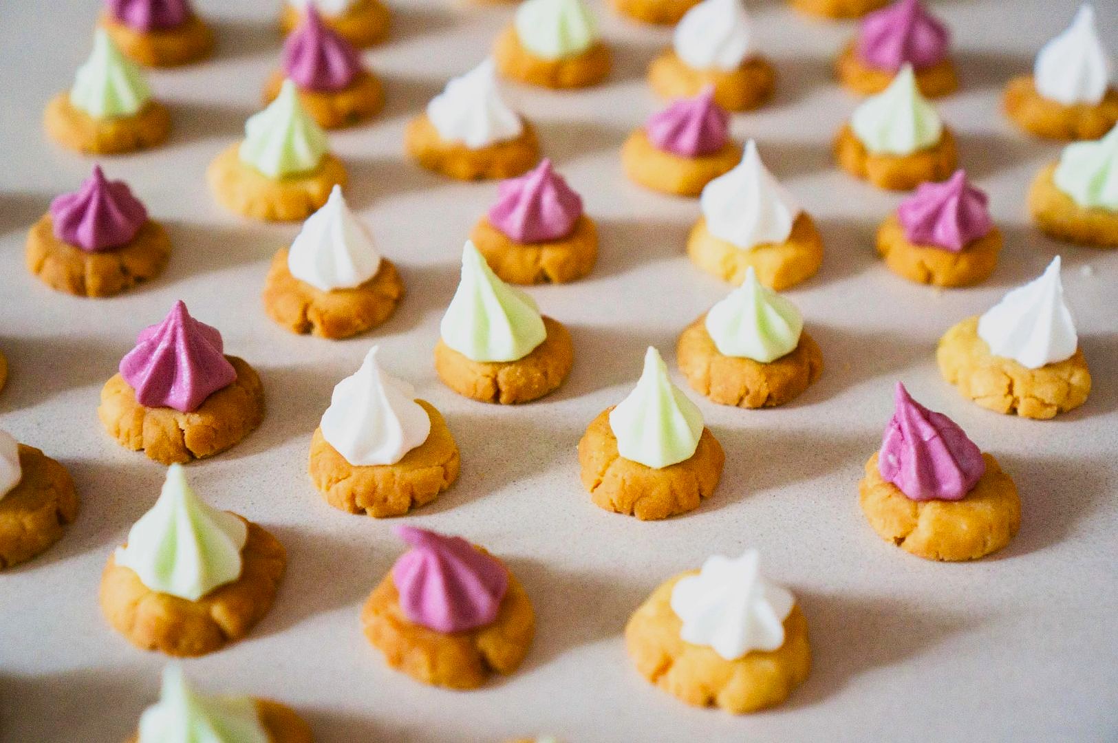Rows of small cookies topped with pastel-colored icing in white, green, and purple on a beige surface, creating a cheerful, playful pattern.