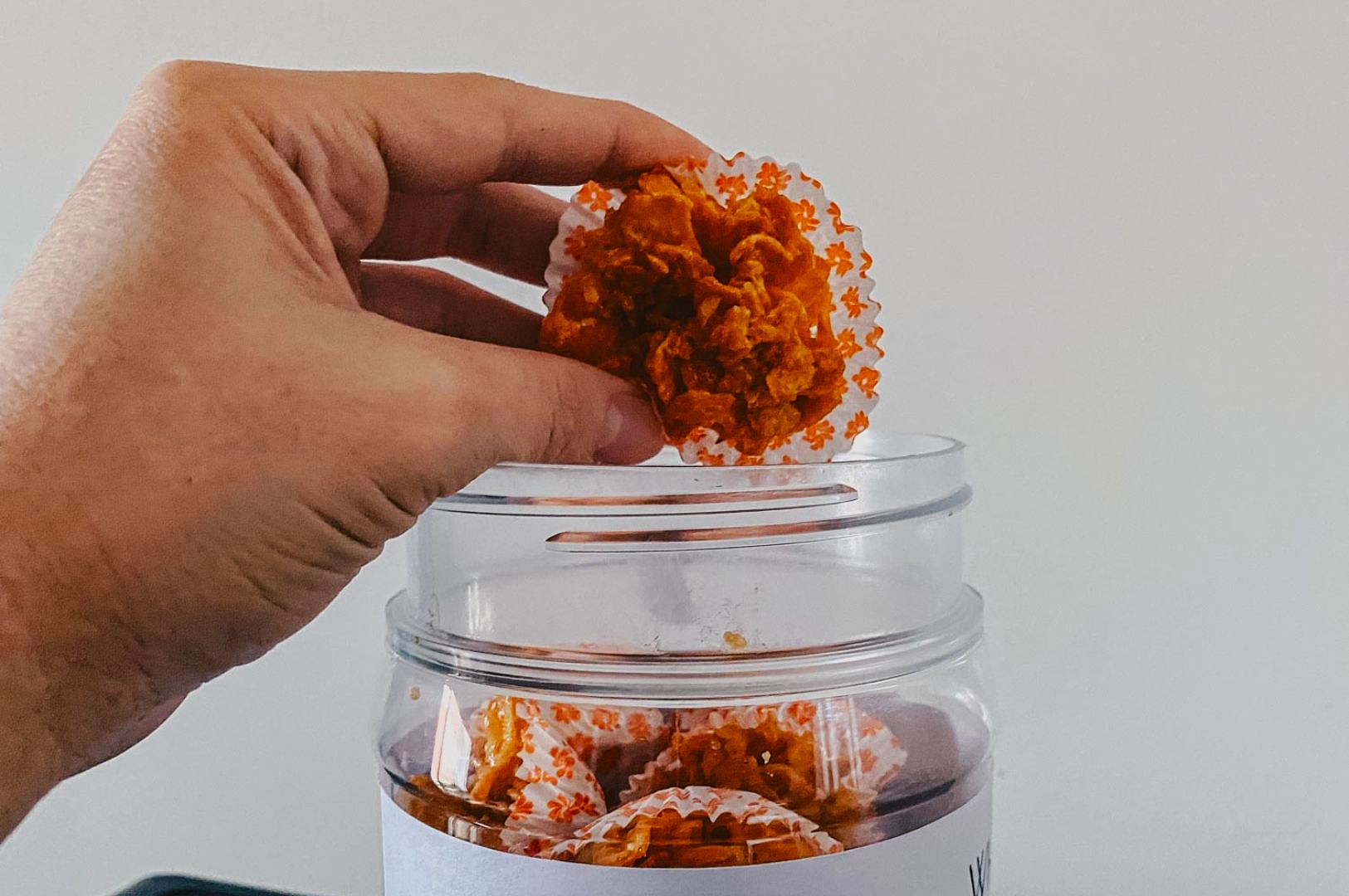 A hand lifts a piece of crunchy, orange snack in a paper liner from a glass jar filled with more snacks. The scene is simple and appetizing.