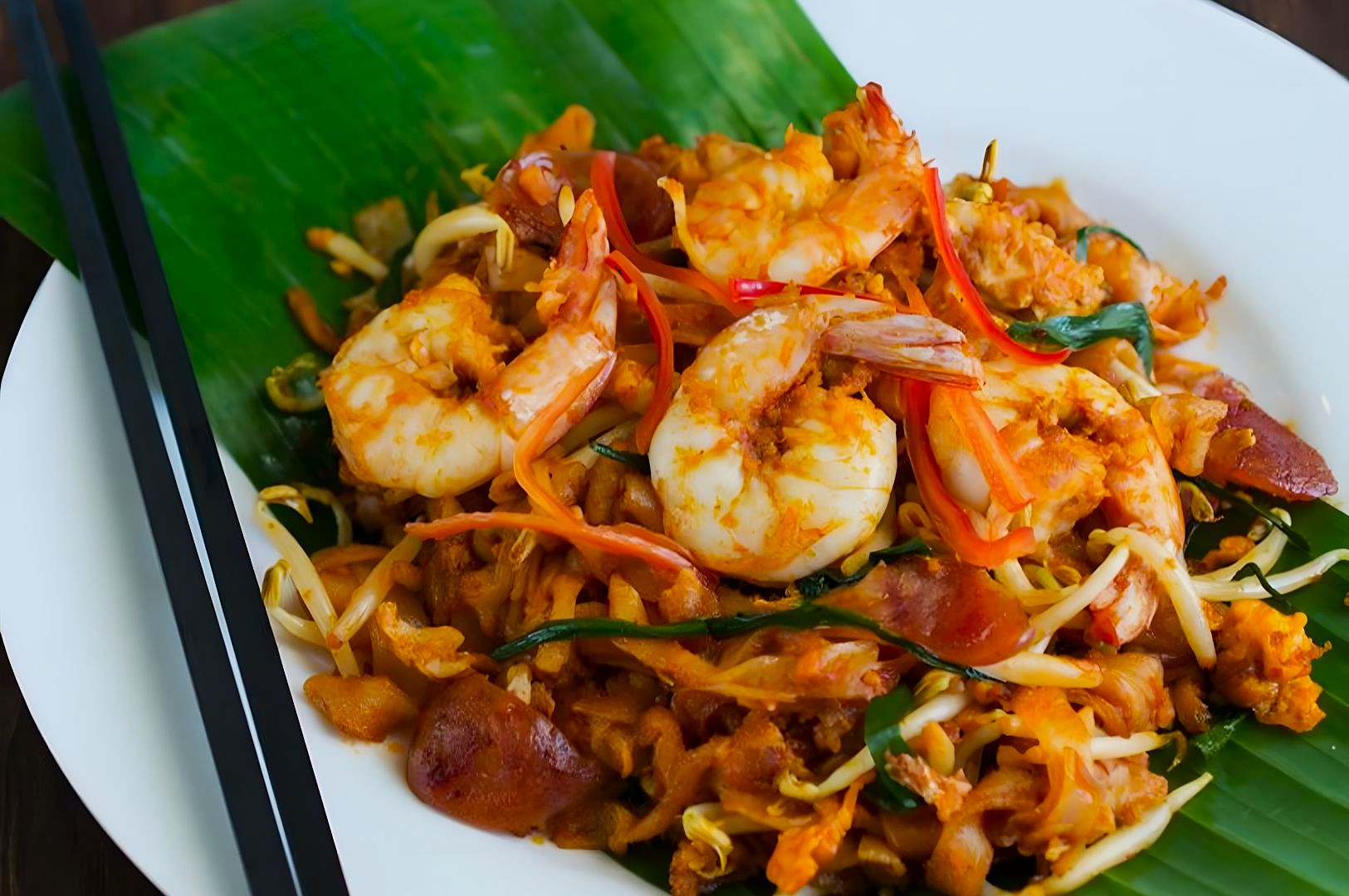 A vibrant dish of Pad Thai with shrimp, bean sprouts, and green onions on a green banana leaf. Black chopsticks rest on the white plate's edge.