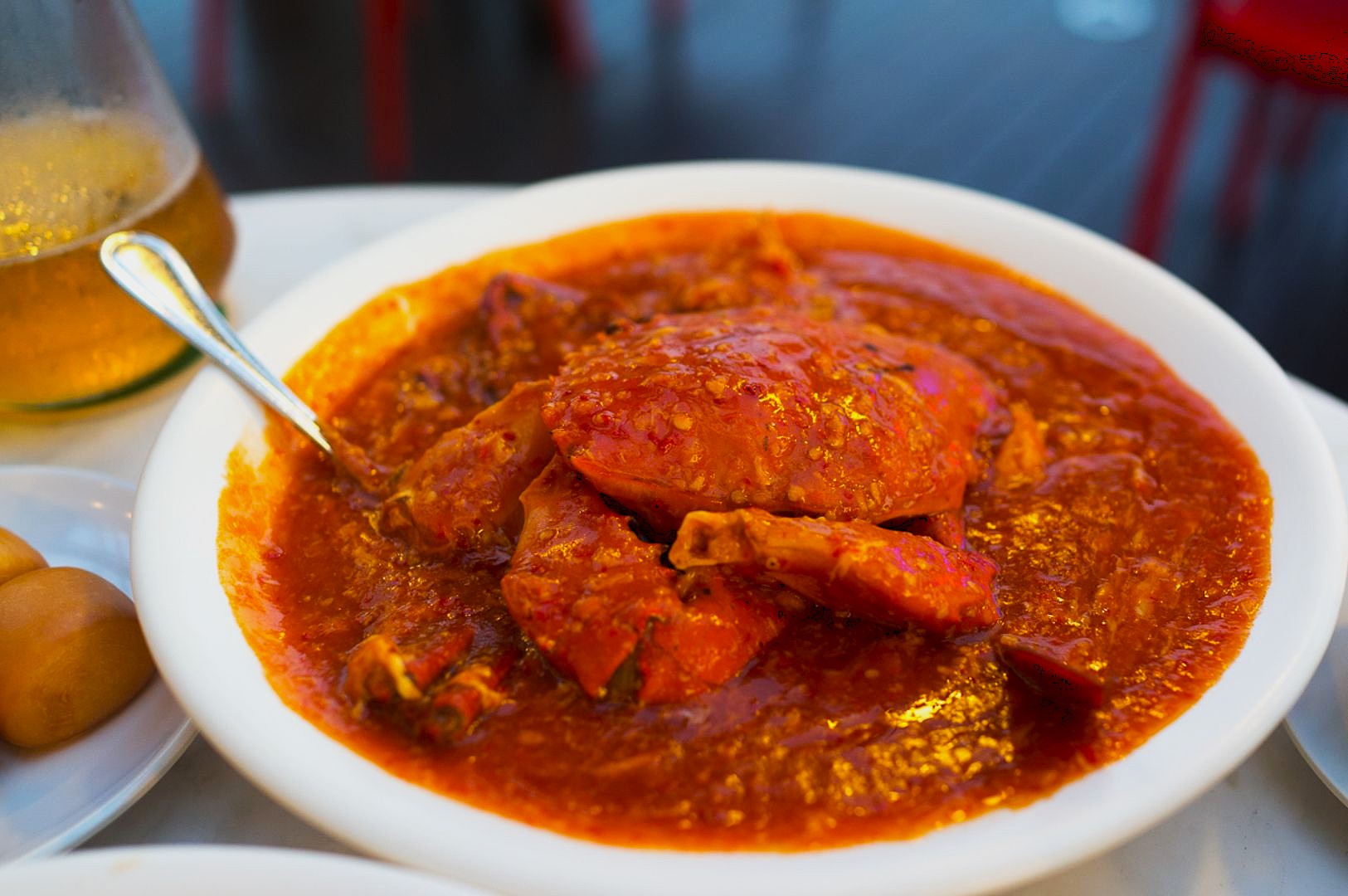 A bowl of chili crab in spicy red sauce with a spoon, set on a white table. The vibrant dish radiates warmth and an inviting aroma.