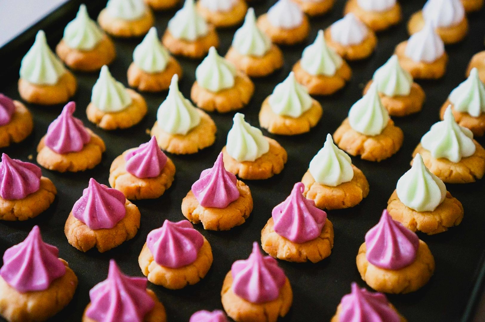 Close-up of neatly arranged cookies topped with colorful meringues in rows on a baking tray. The cookies have pink, white, and light green swirls, creating a playful and festive look.