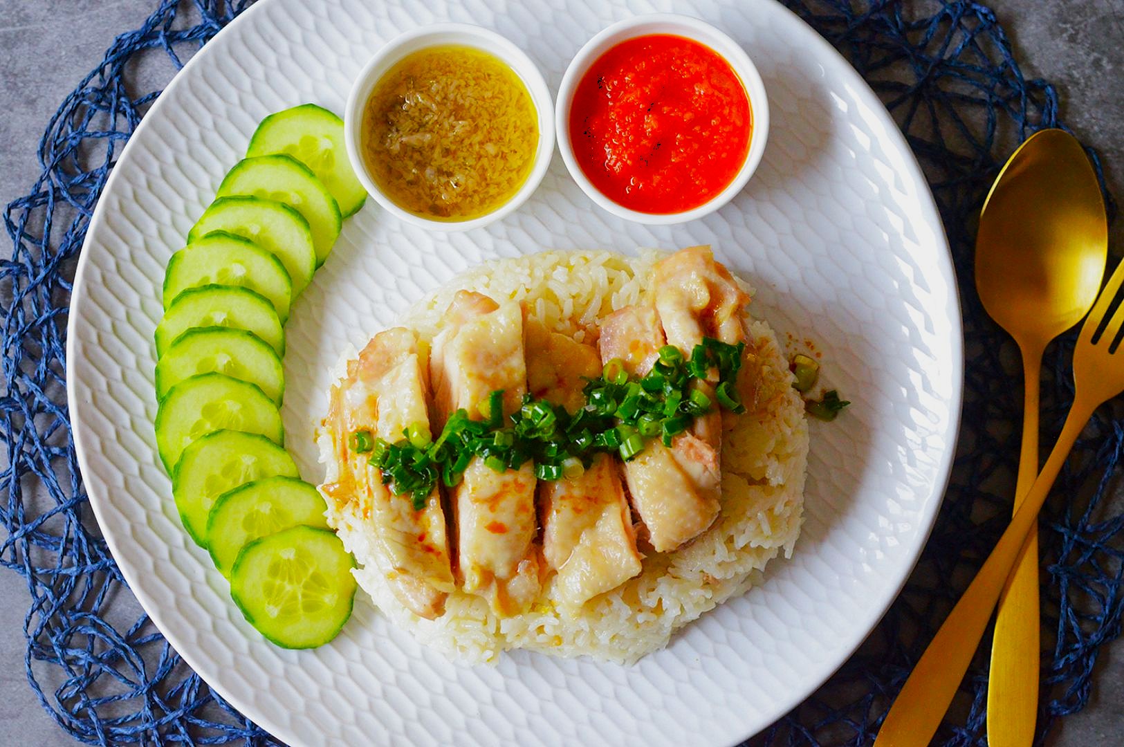 A plate of Hainanese chicken rice garnished with green onions, alongside cucumber slices. Two dipping sauces, red chili and garlic, are on the side. Gold utensils lie nearby.