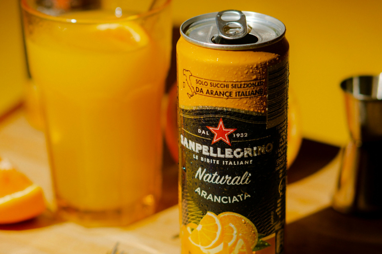 A can of San Pellegrino Aranciata with orange graphics is in focus, with a full glass of orange juice and sliced oranges in a warm, vibrant setting.