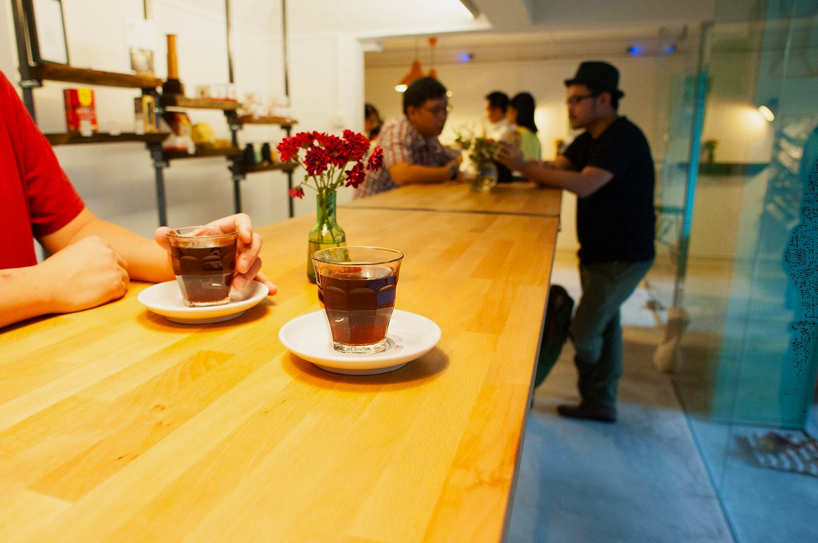 A cozy cafe scene with warm lighting, featuring a wooden counter with two glasses of coffee and red flowers in a vase. Patrons chat in the background.