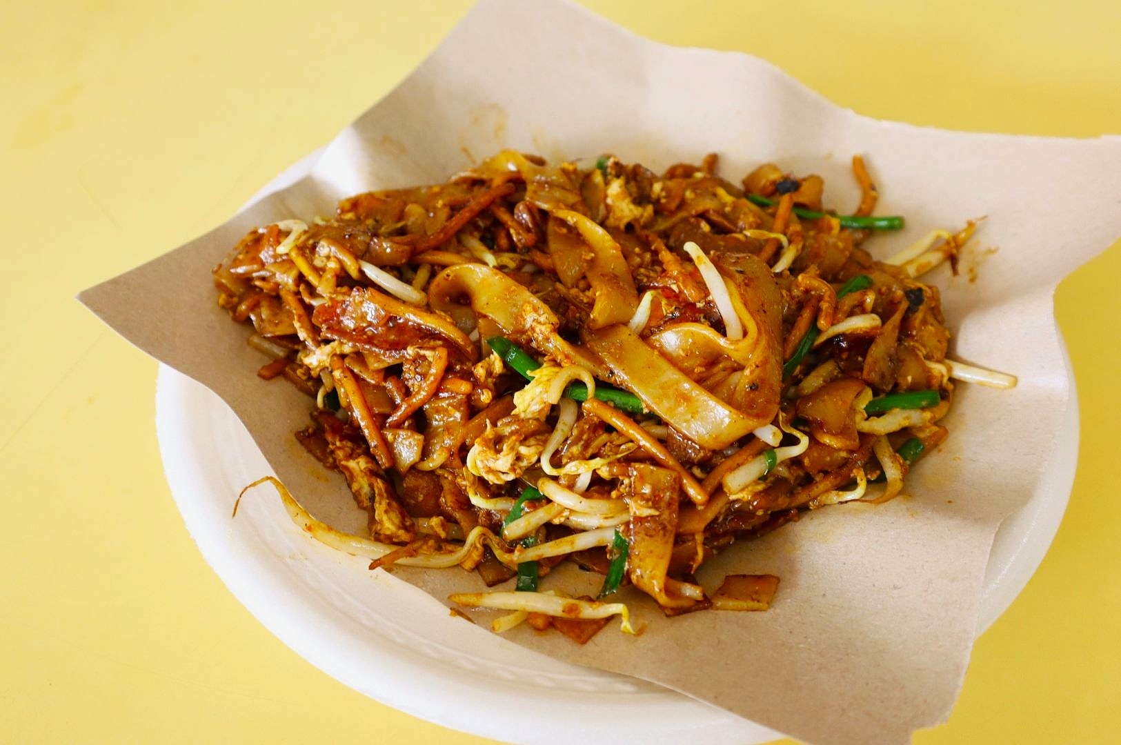 A plate of char kway teow is served on brown paper. The dish features stir-fried flat rice noodles, bean sprouts, and green chives, exuding a savory aroma.