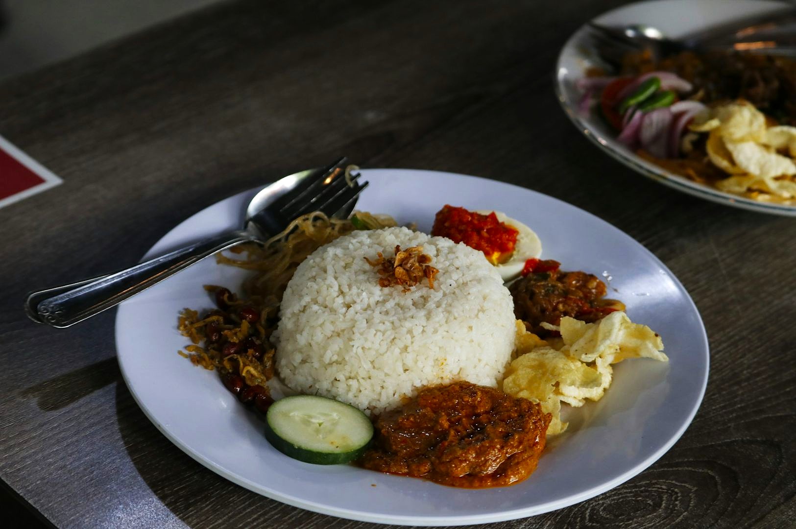 A plate of Indonesian nasi campur on a wooden table, featuring rice topped with fried shallots, surrounded by spicy sambal, beef rendang, crackers, and vegetables.