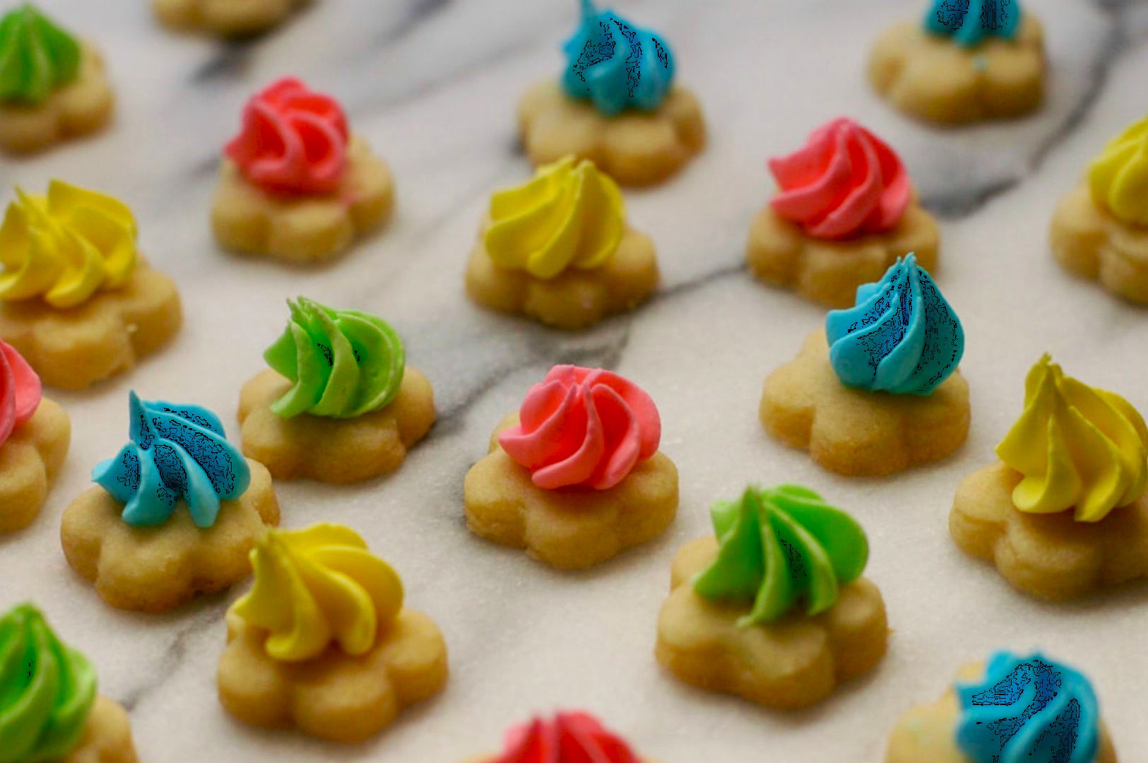 Colorful cookies with pastel icing swirls of pink, blue, yellow, and green are neatly arranged on a marble surface. The tone is playful and joyful.