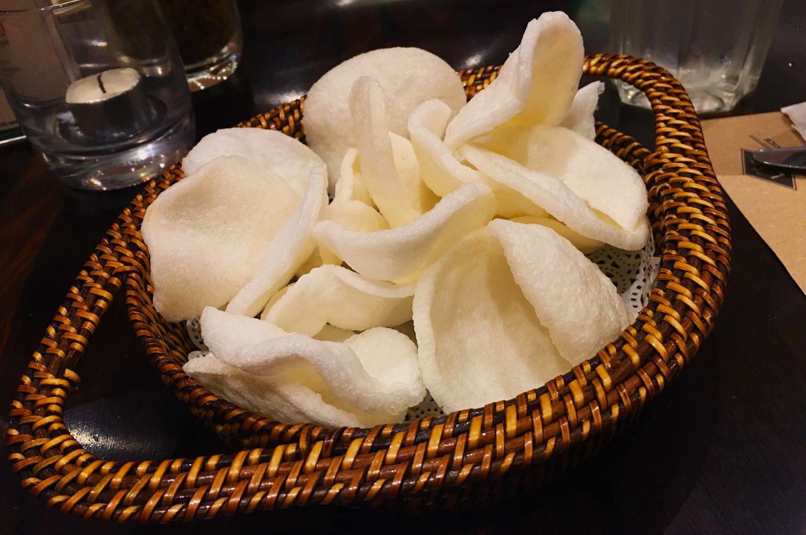 Basket filled with white prawn crackers on a dark wooden table. The basket is woven, and nearby are a glass candle and utensils, creating a cozy dining ambiance.
