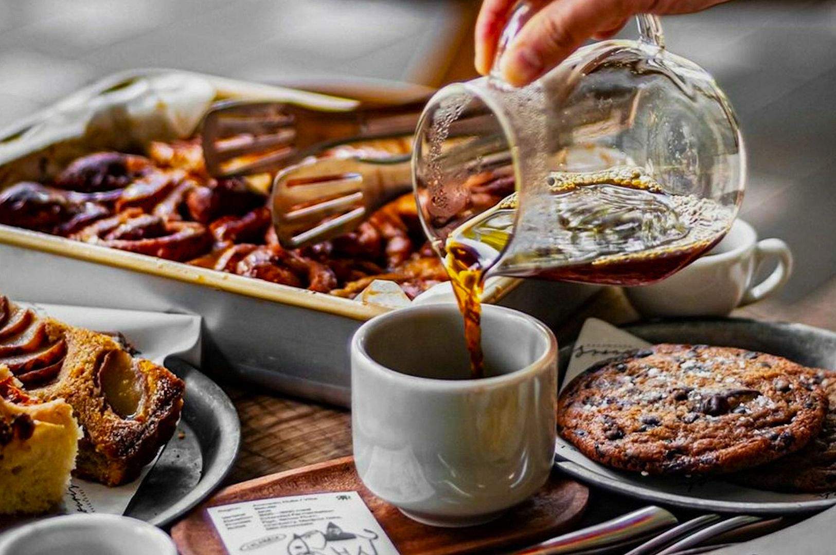 Hand pouring coffee into a cup surrounded by a tray of cinnamon rolls, a chocolate chip cookie, and a slice of cake, creating a cozy and inviting atmosphere.