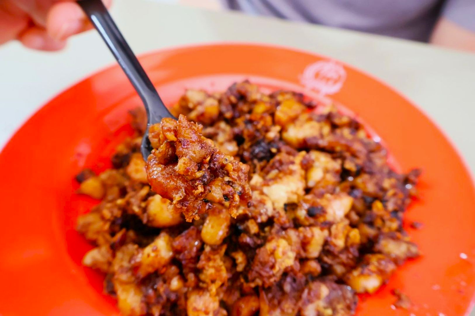 A close-up of a hand holding a black fork with a portion of fried diced radish cake, covered in a savory sauce, on a bright orange plate. The dish looks appetizing and richly seasoned.