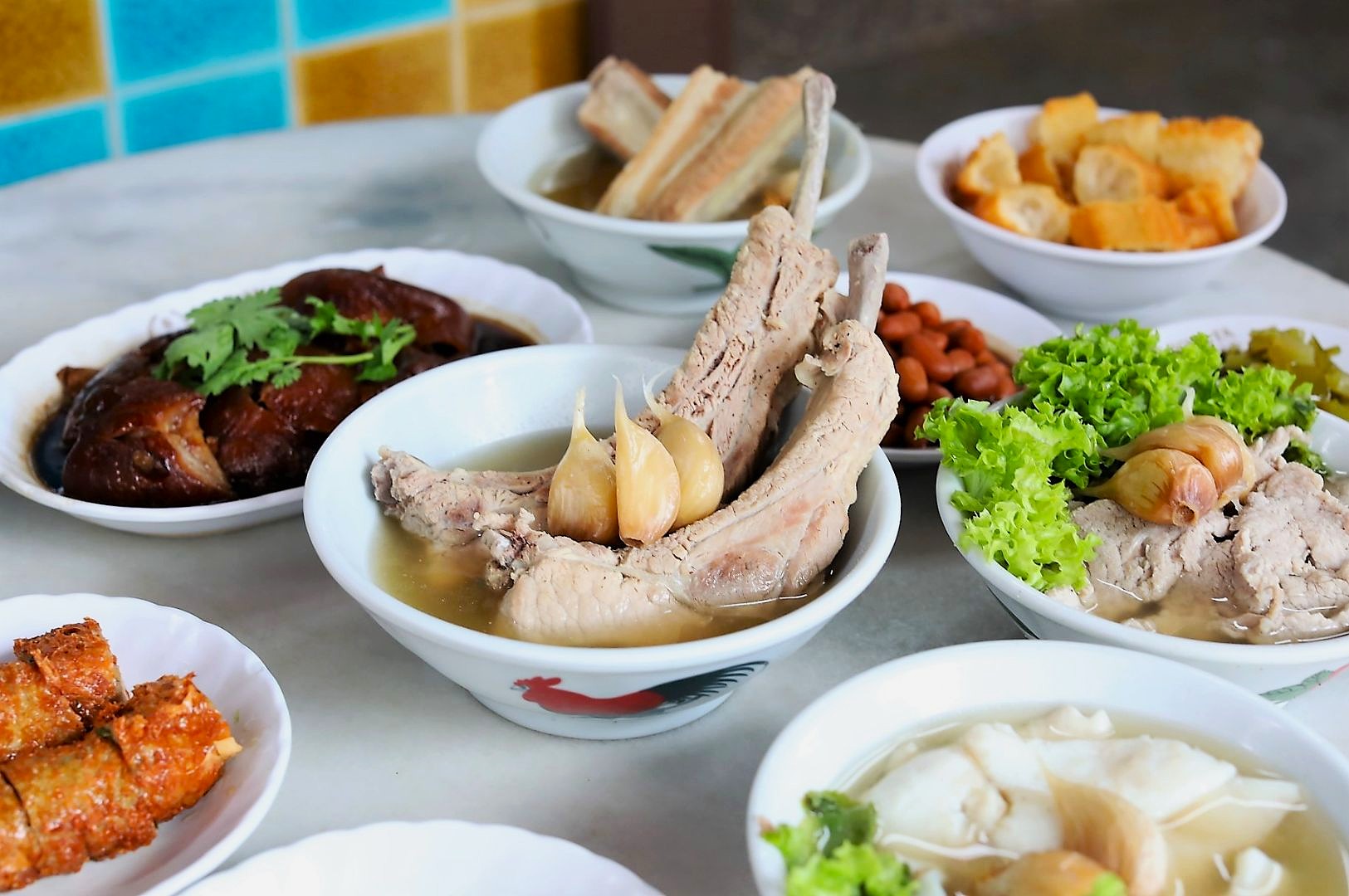 Assorted Asian dishes on a table, featuring pork ribs in broth with garlic, tofu, braised meat, peanuts, and crispy dough, conveying a cozy, communal meal.