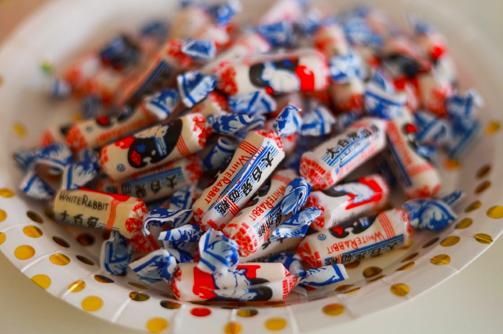 A pile of White Rabbit candy in colorful wrappers, predominantly blue, red, and white, sits in a polka-dotted bowl. The scene evokes nostalgia and sweetness.