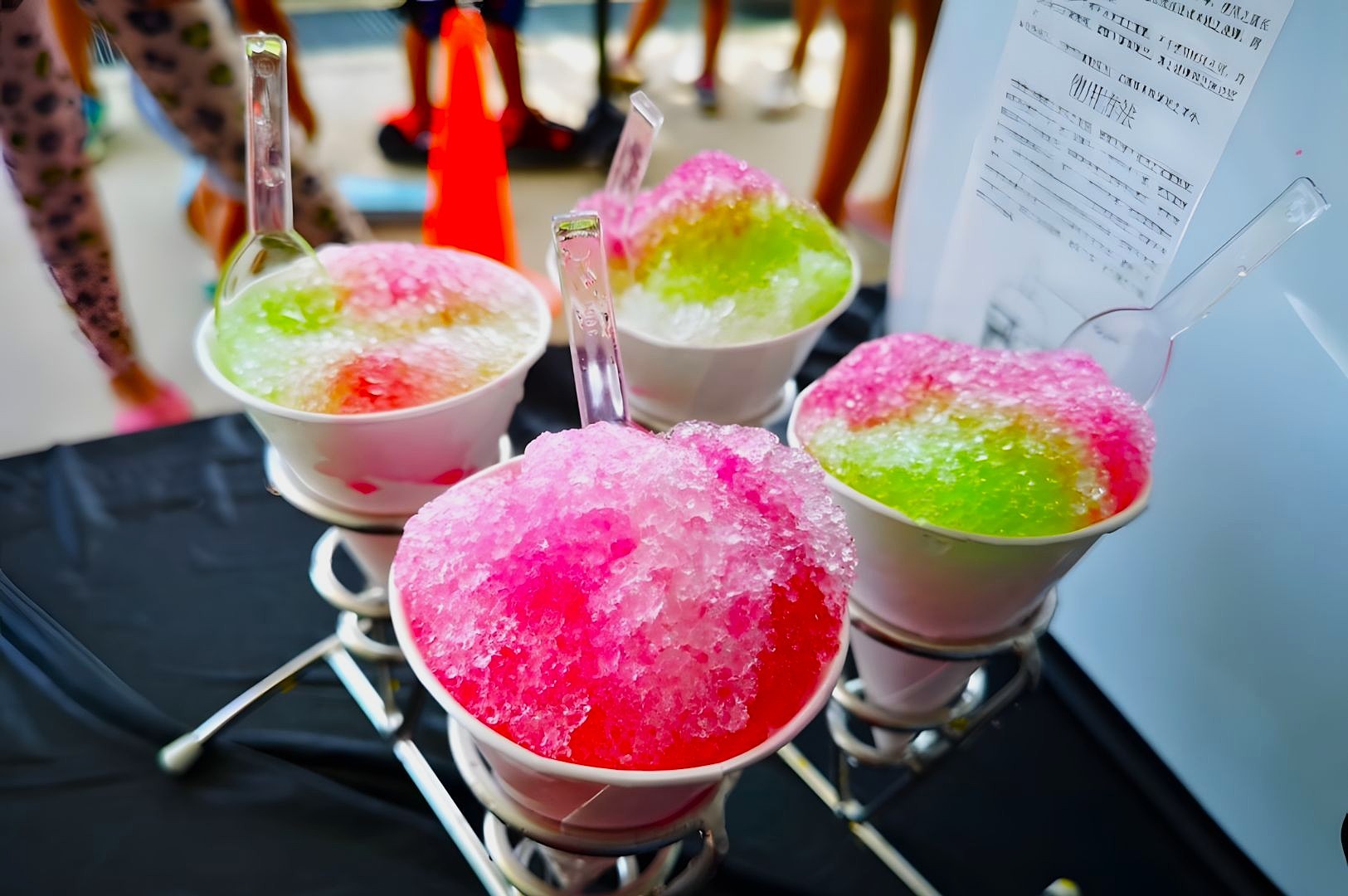 Four cups of colorful shaved ice, green and pink, on a black table with plastic spoons. A cone and legs are visible in the blurred background.