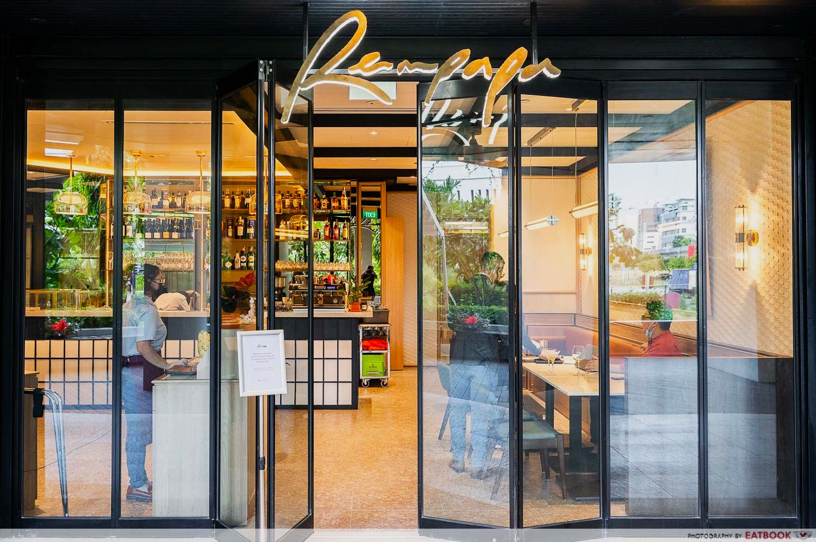 Elegant restaurant entrance with large glass doors and soft lighting. Inside, a stocked bar is visible. Few diners sit at tables in a cozy setting.