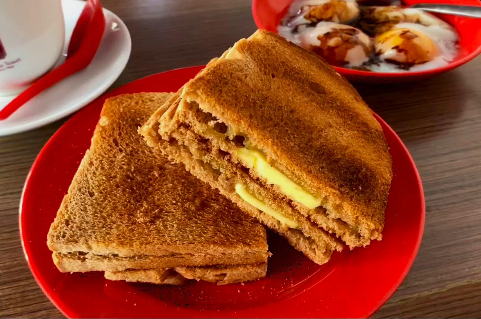 Toasted bread with butter on a red plate, next to soft-boiled eggs drizzled with soy sauce in the background. Warm, cozy breakfast setting.