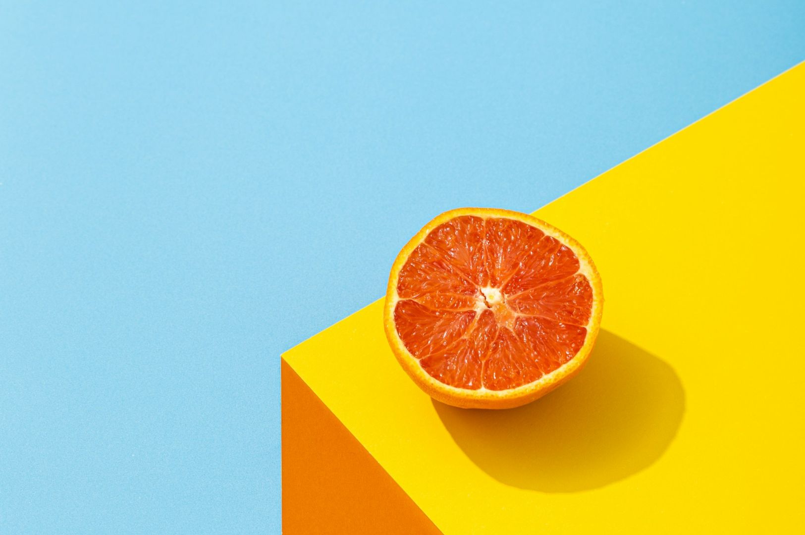 Half an orange sits on a yellow surface against a blue background, casting a shadow. The bright colors create a vibrant and fresh atmosphere.