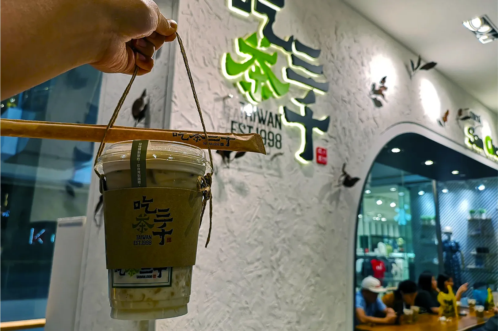 A hand holds a bubble tea cup with a wooden carrier in a modern cafe. The cafe's textured wall features Asian characters and a neon sign. The atmosphere is lively.