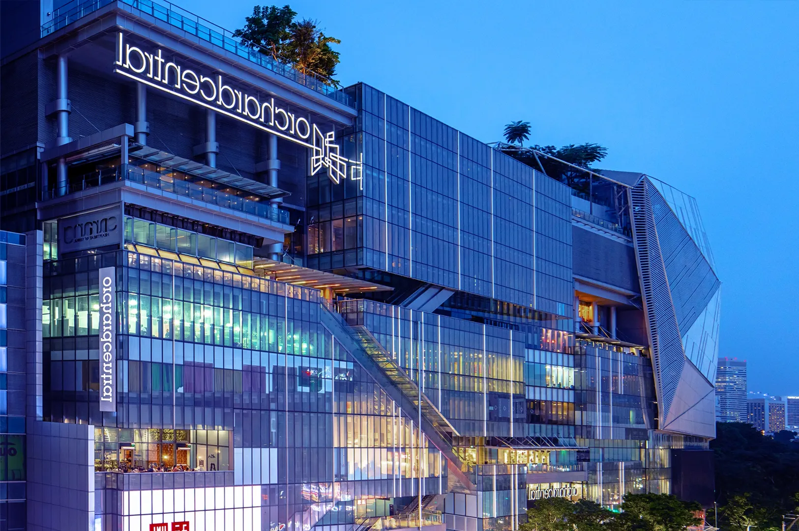 Modern glass building at dusk, featuring bright, colorful lights and sleek design. Urban and futuristic ambiance with trees on the rooftop.