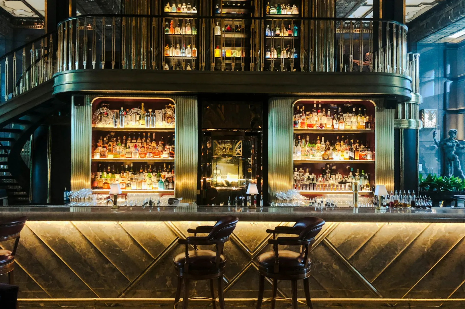 Luxurious bar with a marble counter, high-backed chairs, and warm lighting. Bottles line tall shelves behind, surrounded by elegant gold accents.
