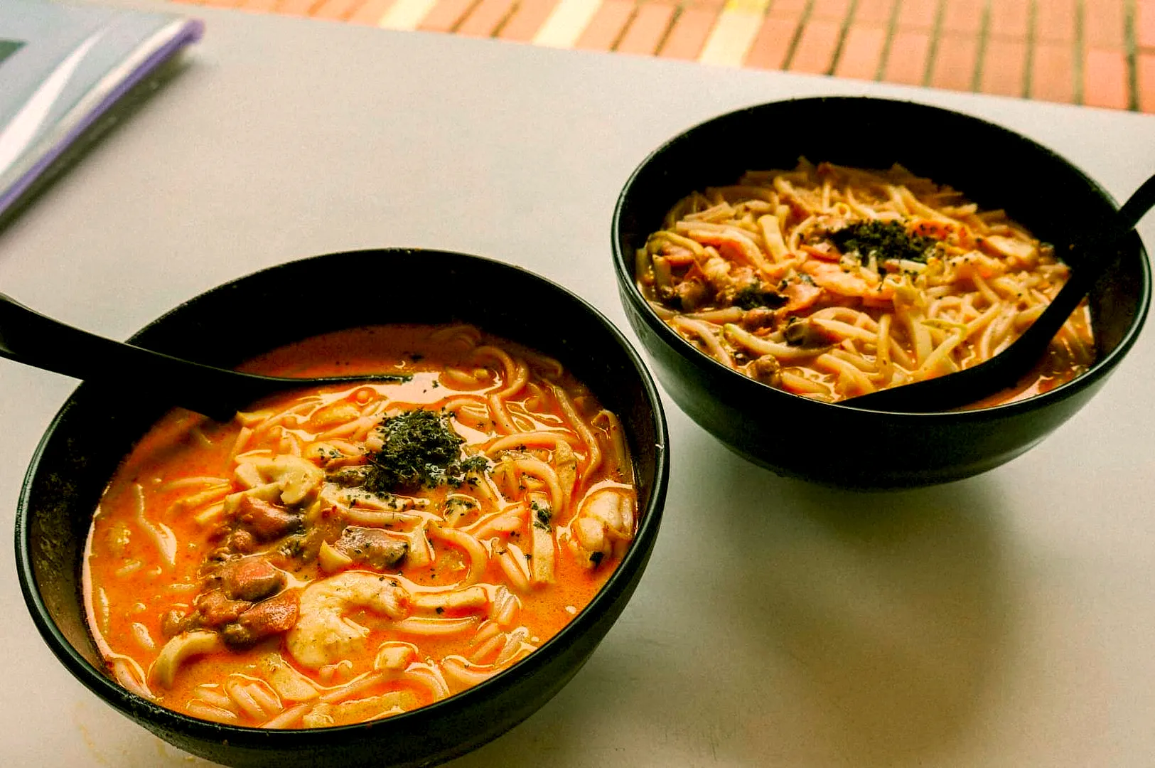 Two black bowls of ramen on a light table, filled with noodles, shrimp, and broth, garnished with herbs. A cozy, inviting meal setting.