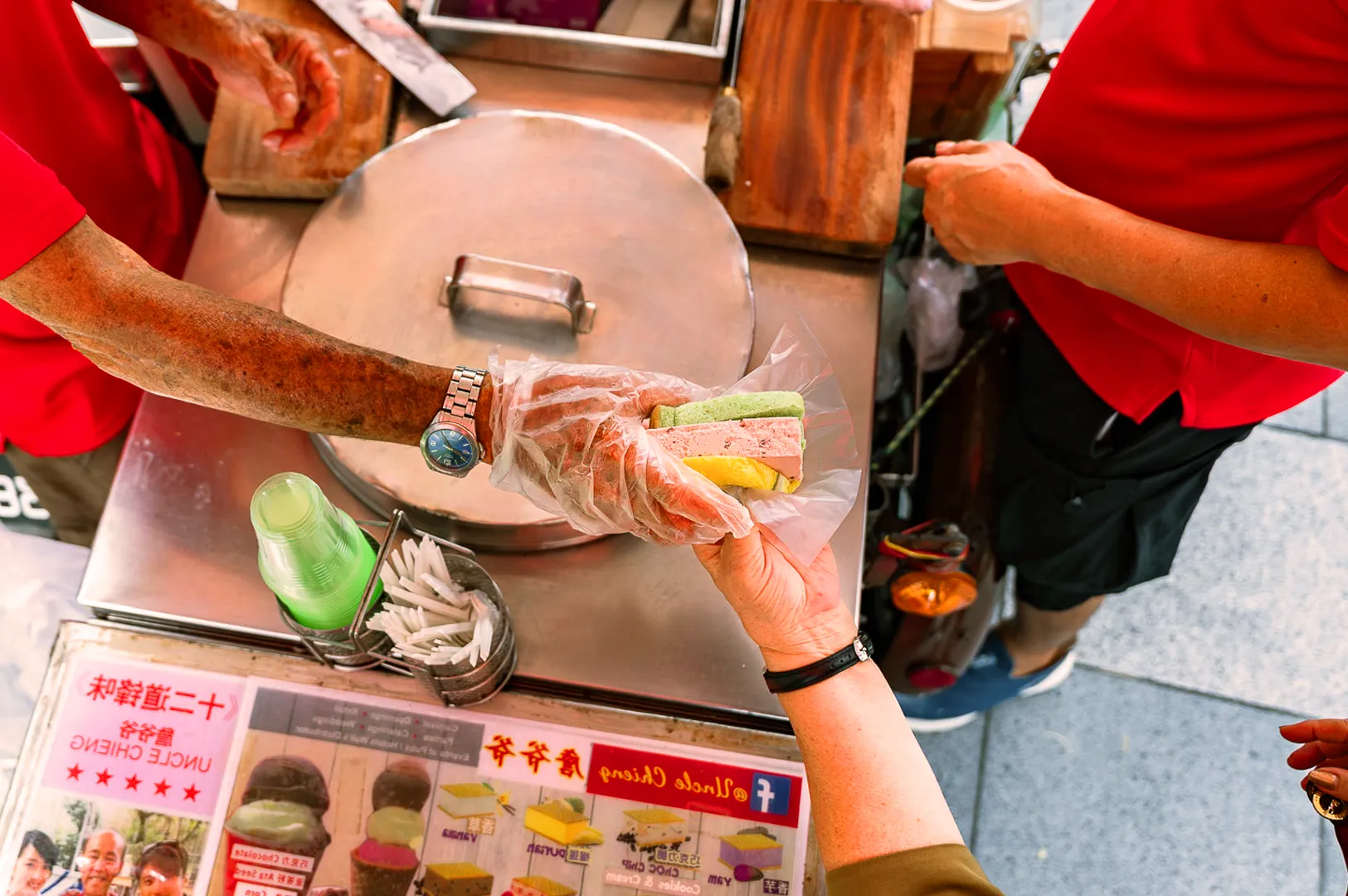 A street vendor in a red shirt hands an ice cream sandwich in rainbow-colored bread to a customer. The setting is vibrant and bustling.