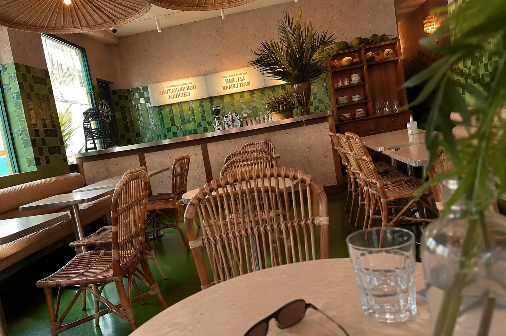 Modern cafe interior featuring woven rattan chairs, green mosaic tiled walls, tropical palm plants, and wooden shelving creating a relaxed Singapore dining atmosphere