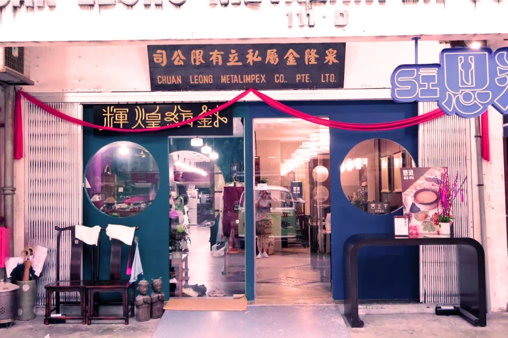 Shopfront with circular blue doors, a red ribbon, and Chinese characters. Warm interior lighting. Sign reads "Chuan Leong Metalimpex Co. Pte. Ltd."