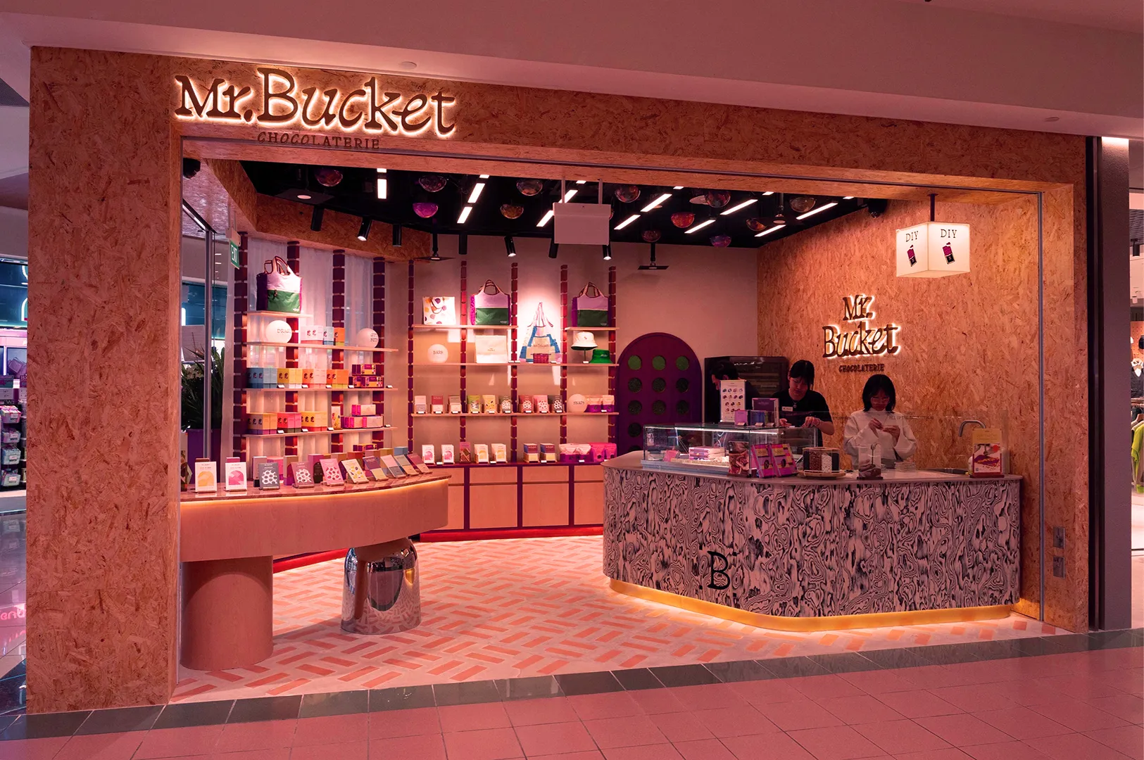 Storefront of Mr. Bucket Chocolaterie with colorful packaging on shelves, two staff behind a patterned counter. Warm, welcoming ambiance.