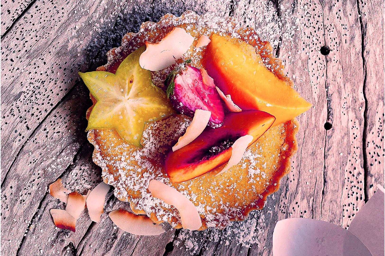 A tart topped with star fruit, peaches, and coconut shavings on a rustic wooden surface. The tart is dusted with powdered sugar, giving a fresh, vibrant look.