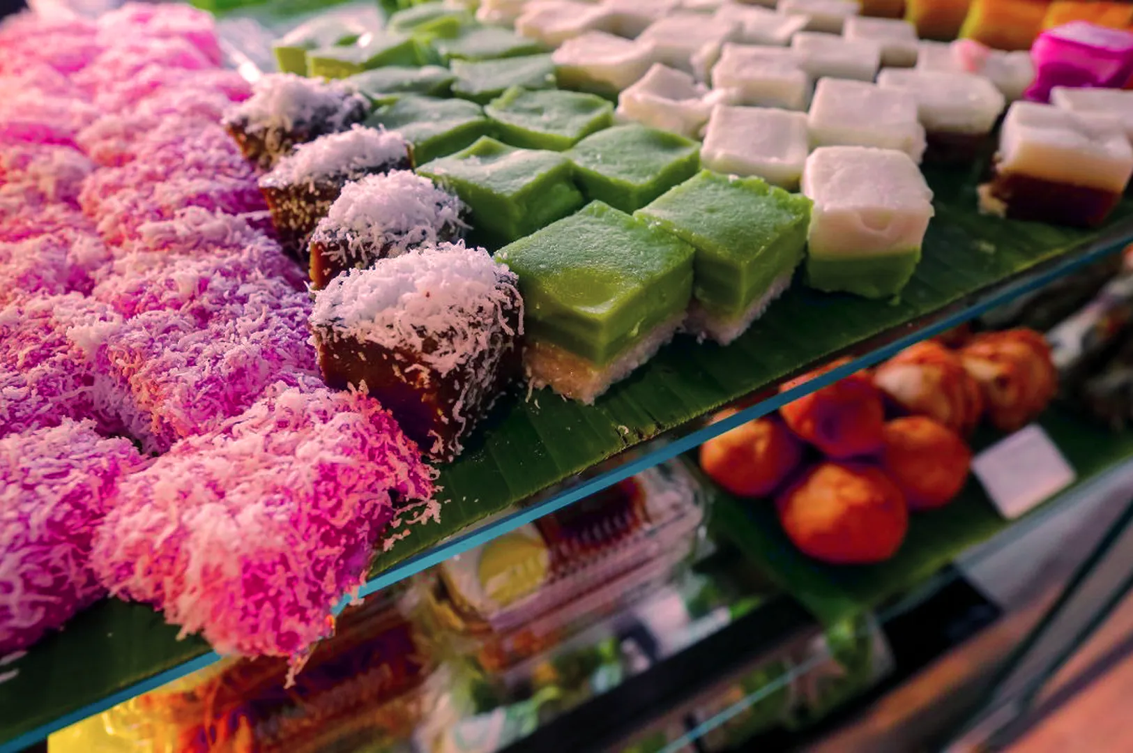 A display of colorful Malaysian kuih on banana leaves, featuring pink, coconut-covered desserts and layered green squares. A warm, inviting market scene.