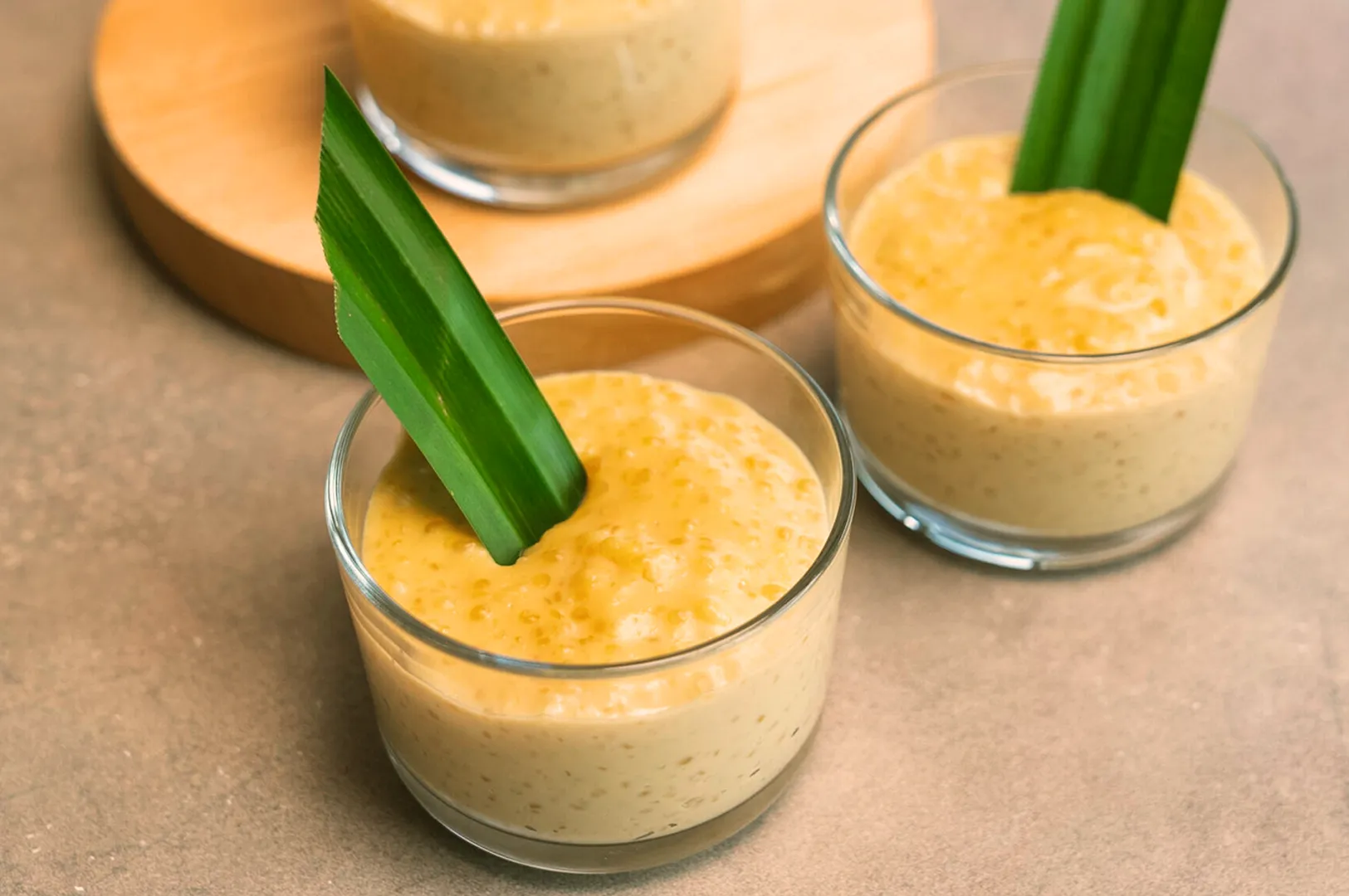 Three clear glasses filled with creamy pudding are topped with green leaves. They sit on a light brown surface, exuding a fresh and tropical vibe.