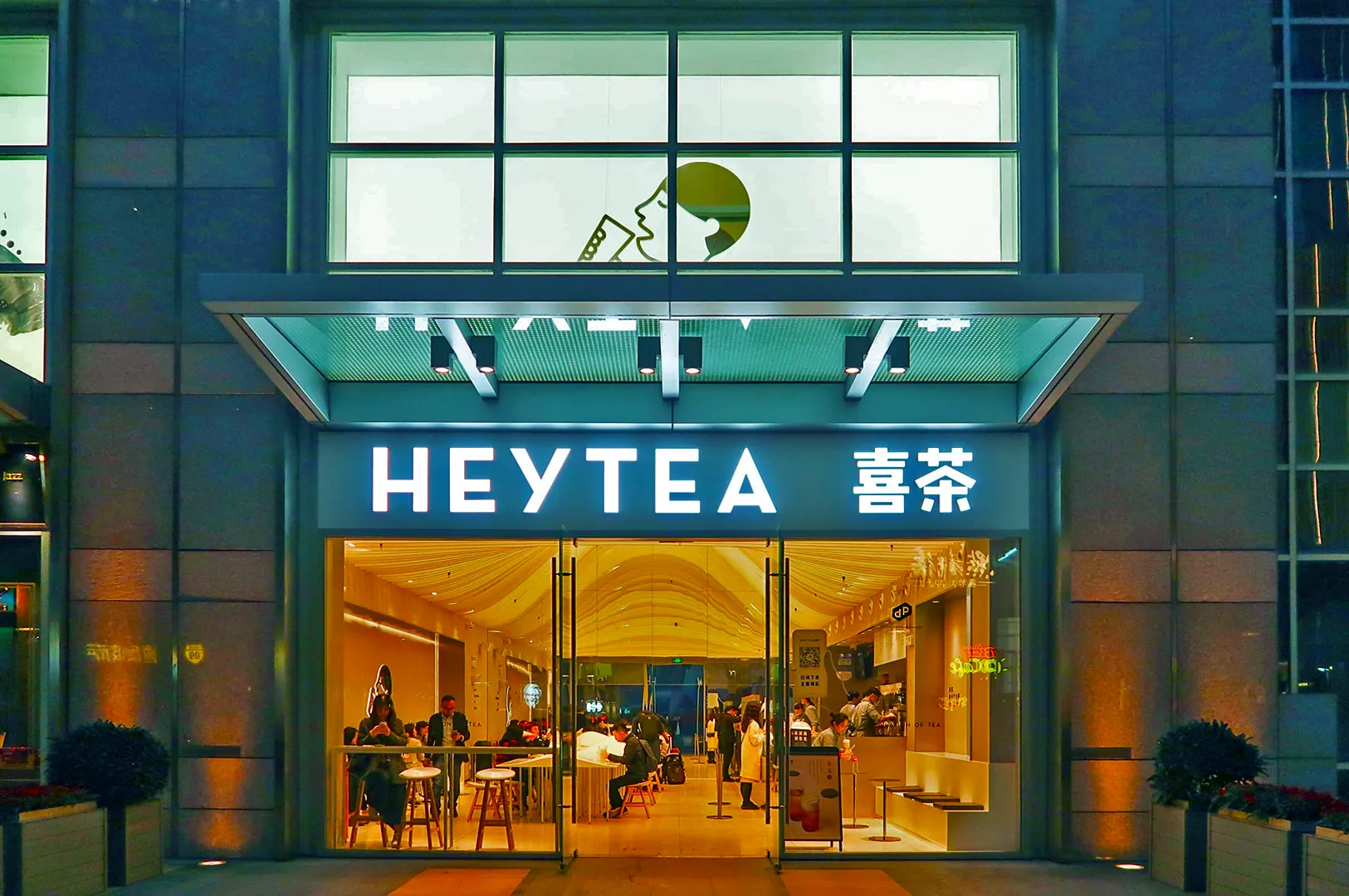 Exterior view of a Heytea store at night, with bright signage and large glass entrance. Inside, customers sit and stand, creating a lively atmosphere.