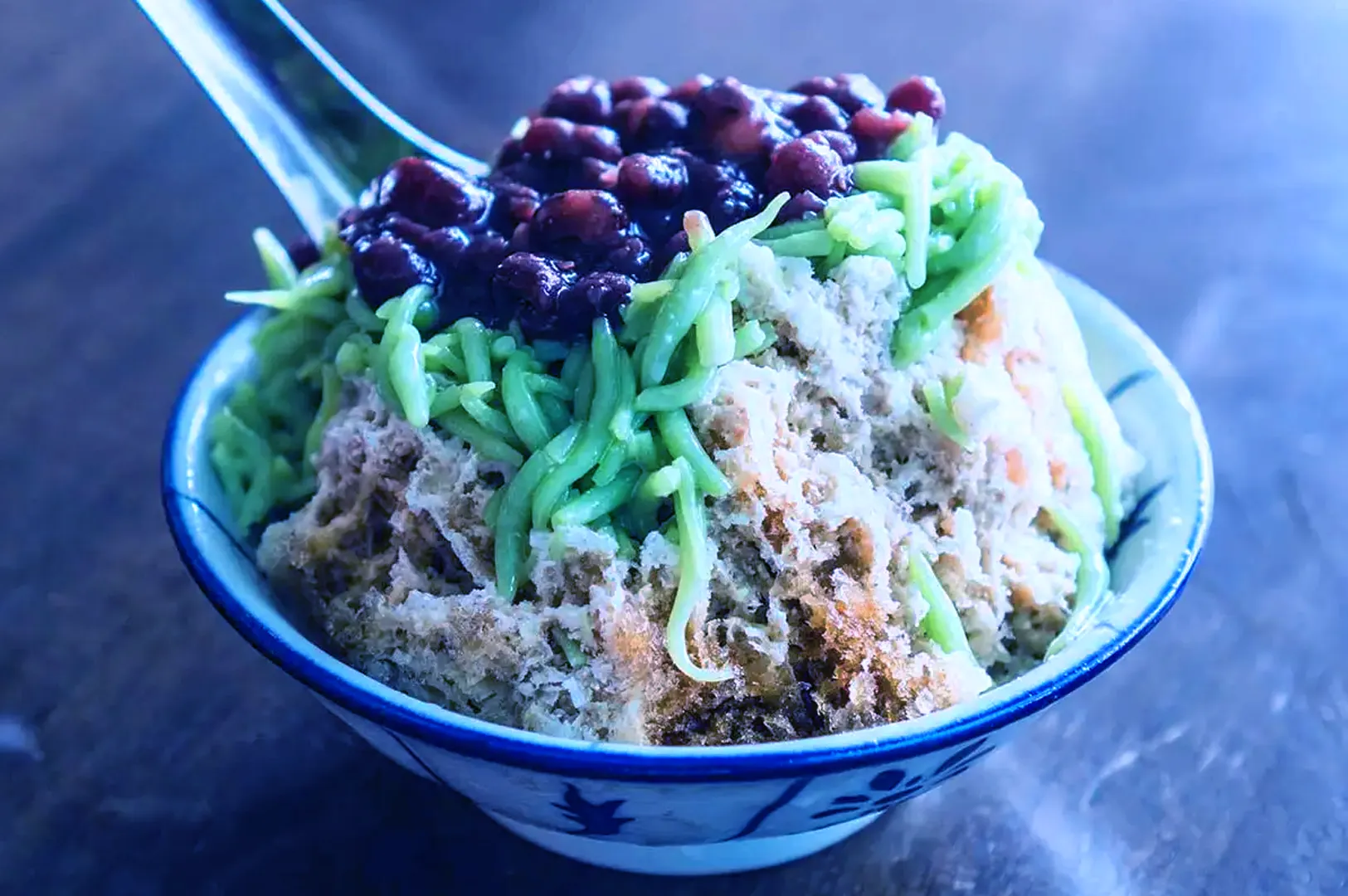 A bowl of cendol on a table, featuring shaved ice topped with green pandan noodles and red beans, creating a refreshing dessert appearance.