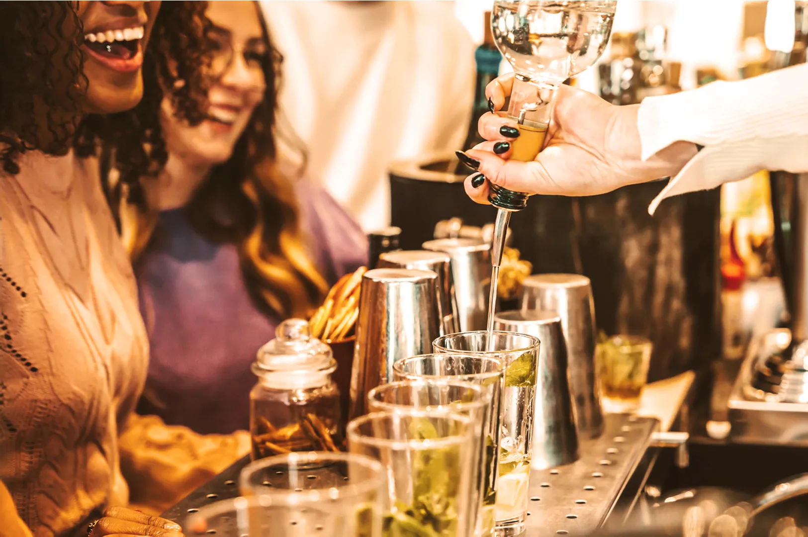 A joyful scene at a bar with laughing patrons. A hand pours clear liquid into glasses with mint. Warm, lively atmosphere with cocktail shakers visible.