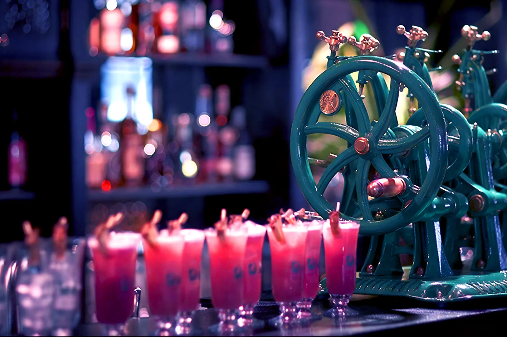Rows of vibrant pink cocktails line a bar counter, set beside vintage green machinery. The warm, colorful scene feels lively and inviting.
