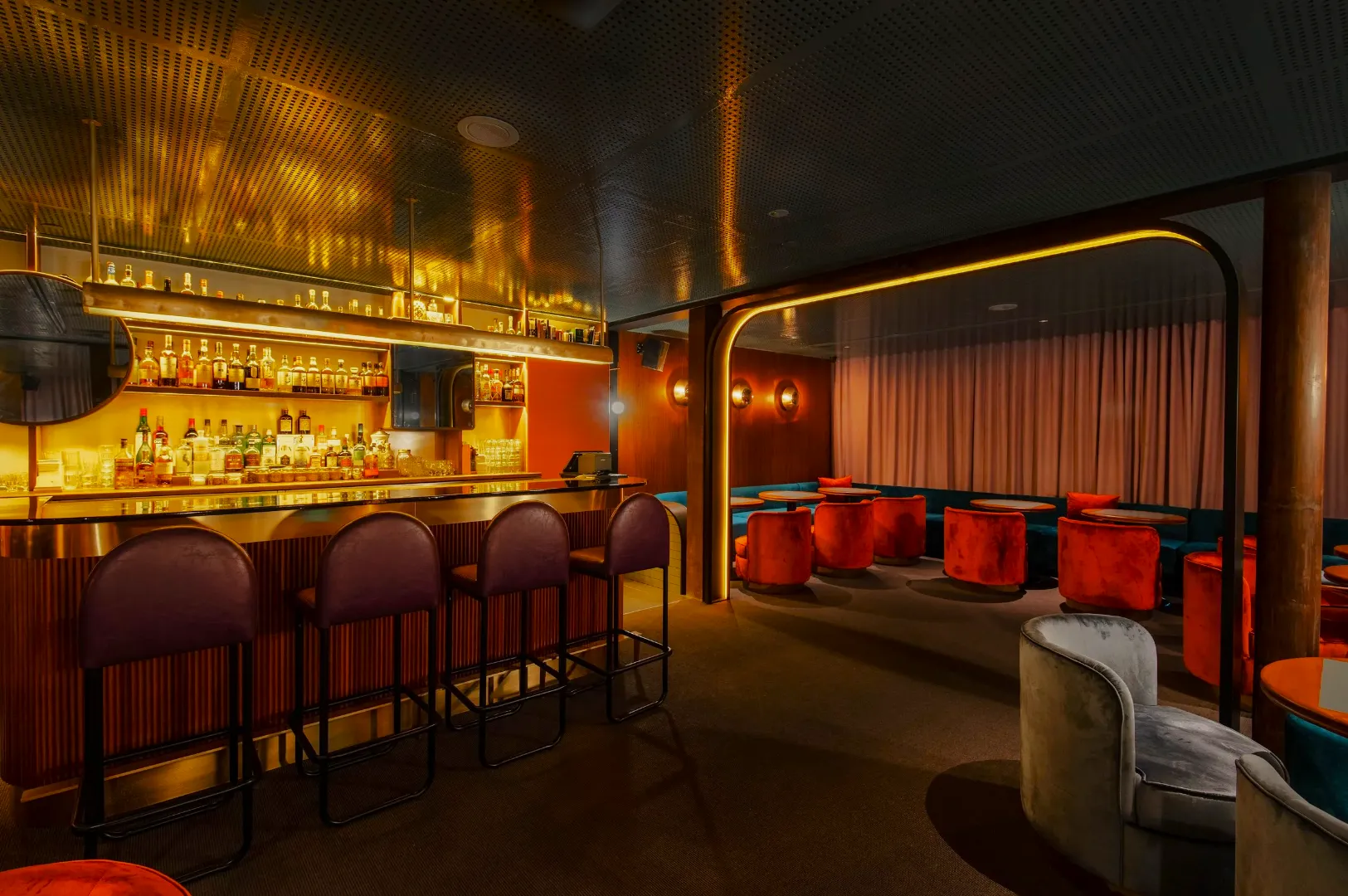 Chic bar interior with dim lighting, featuring a stocked bar with high stools on the left and plush velvet seating in orange and gray on the right, creating a cozy ambiance.