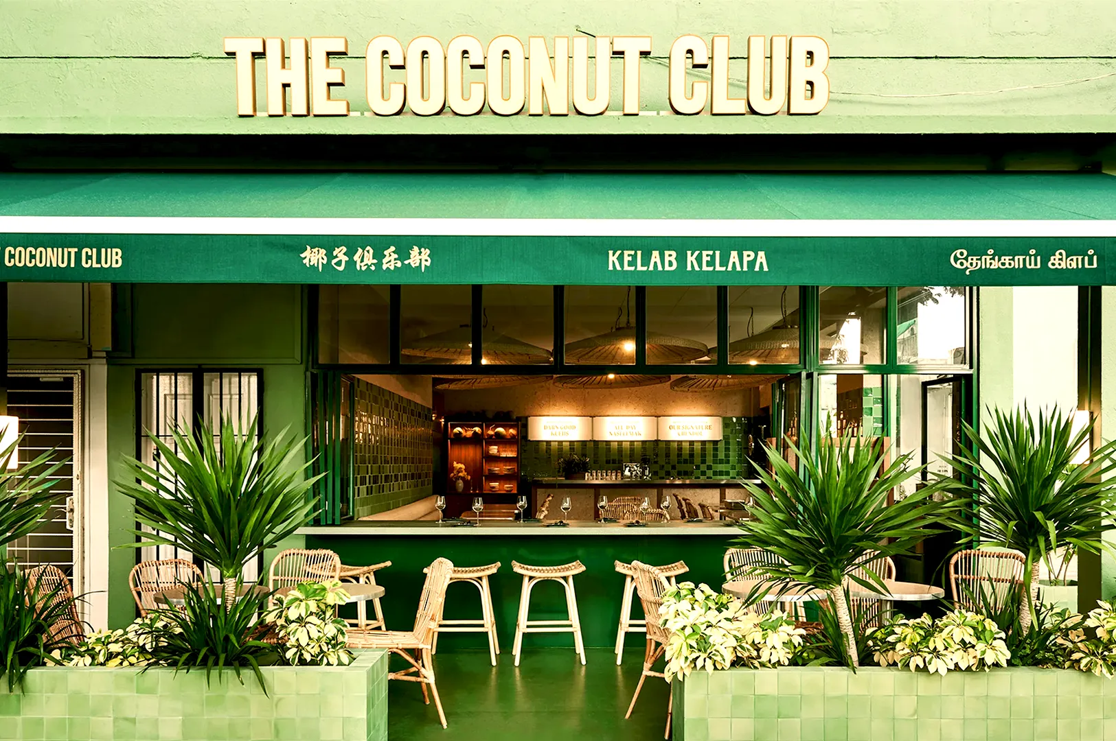 Front view of "The Coconut Club" cafe with a green facade, tropical plants, and wicker bar stools. The atmosphere is inviting and vibrant.