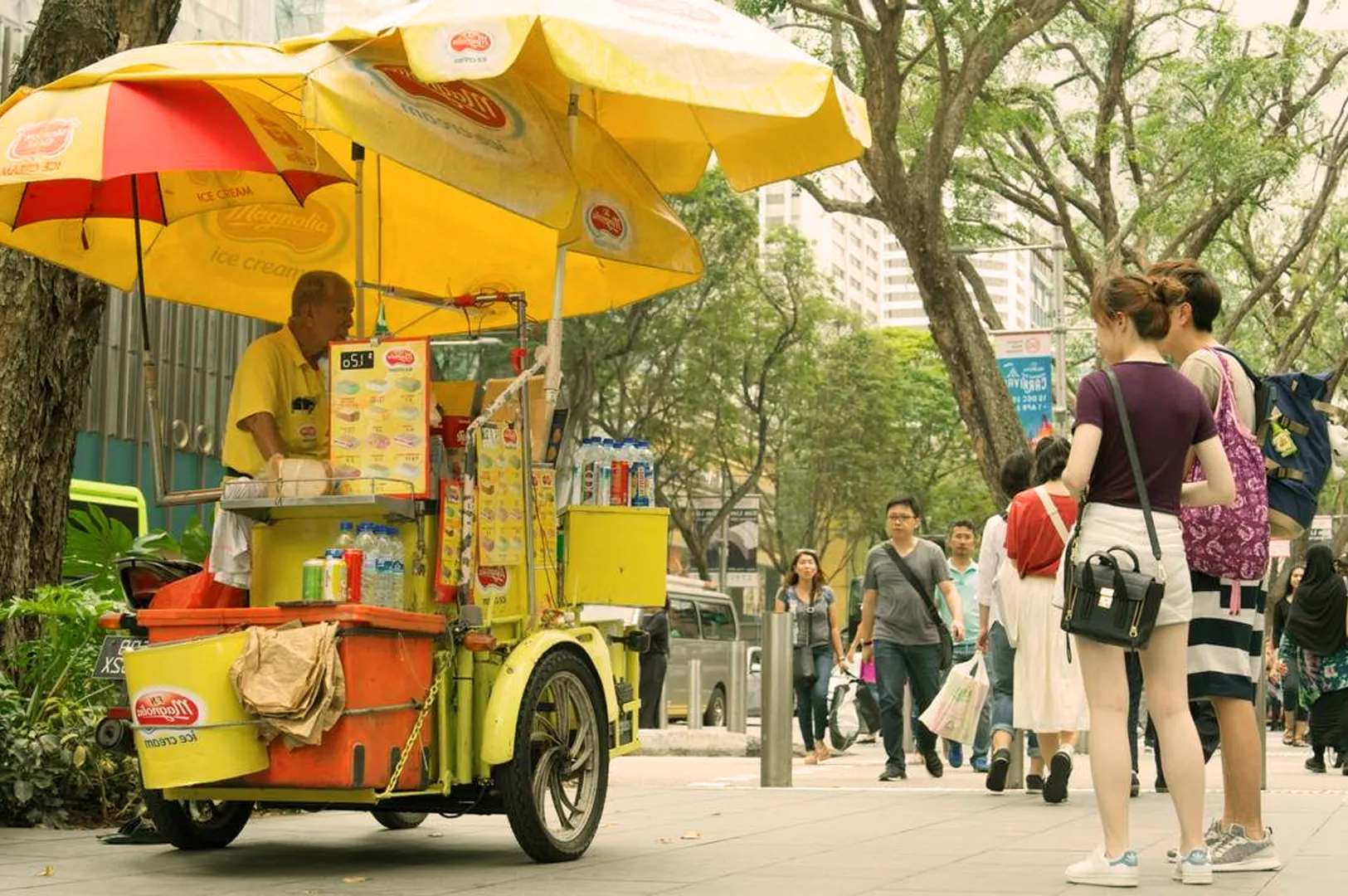 A vibrant street scene shows an ice cream vendor in a yellow cart with red and yellow umbrellas. Two people are ordering, while others walk nearby. The mood is lively.