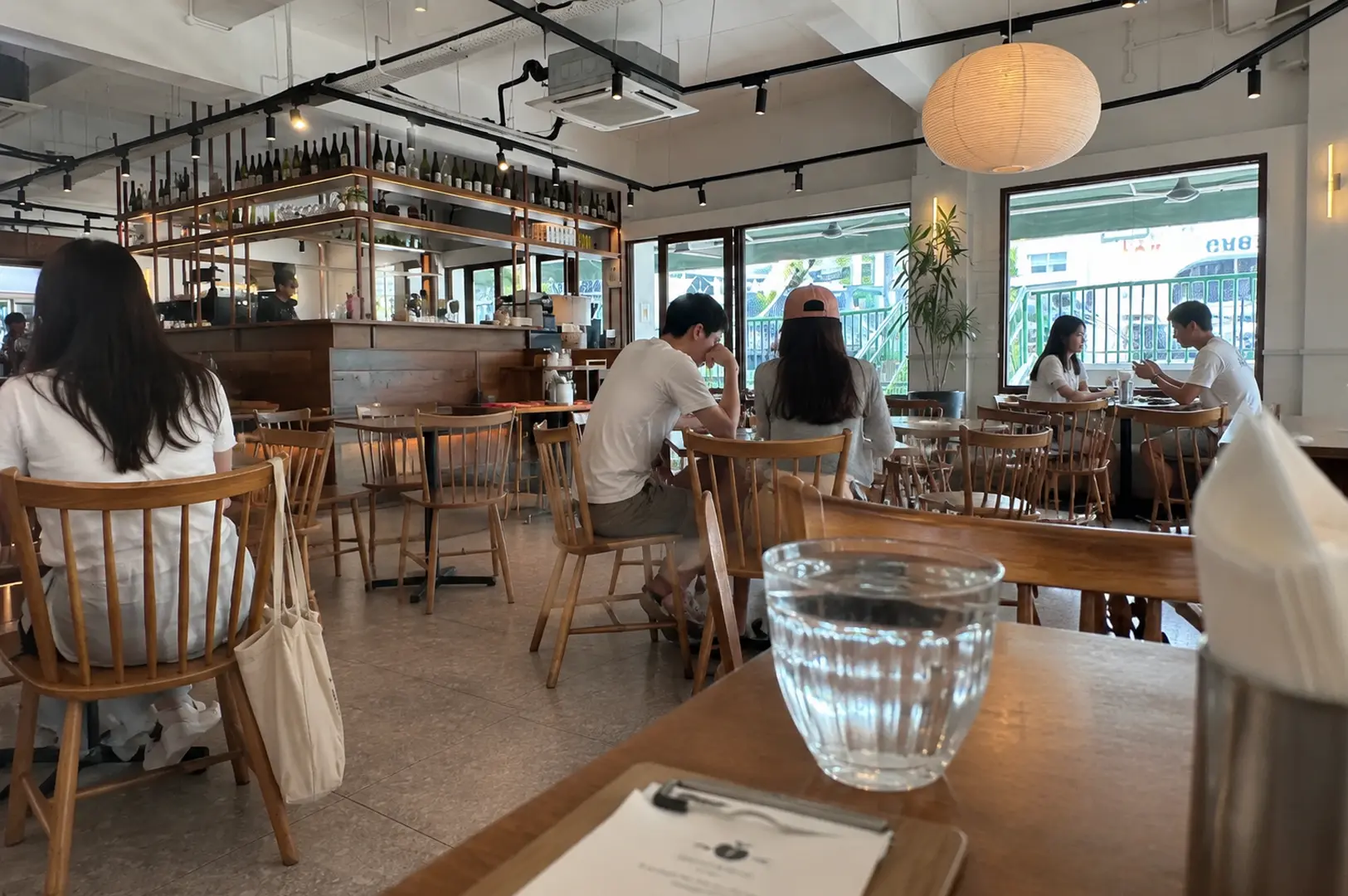 Bright modern cafe with wooden Windsor chairs, customers dining at tables, wine bottles displayed on metal shelving at the bar, large paper lantern pendant light, and floor-to-ceiling windows