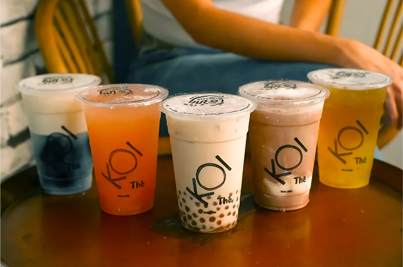 Five bubble tea drinks on a wooden table, each a different color, with a person sitting behind. The setting is cozy and inviting.