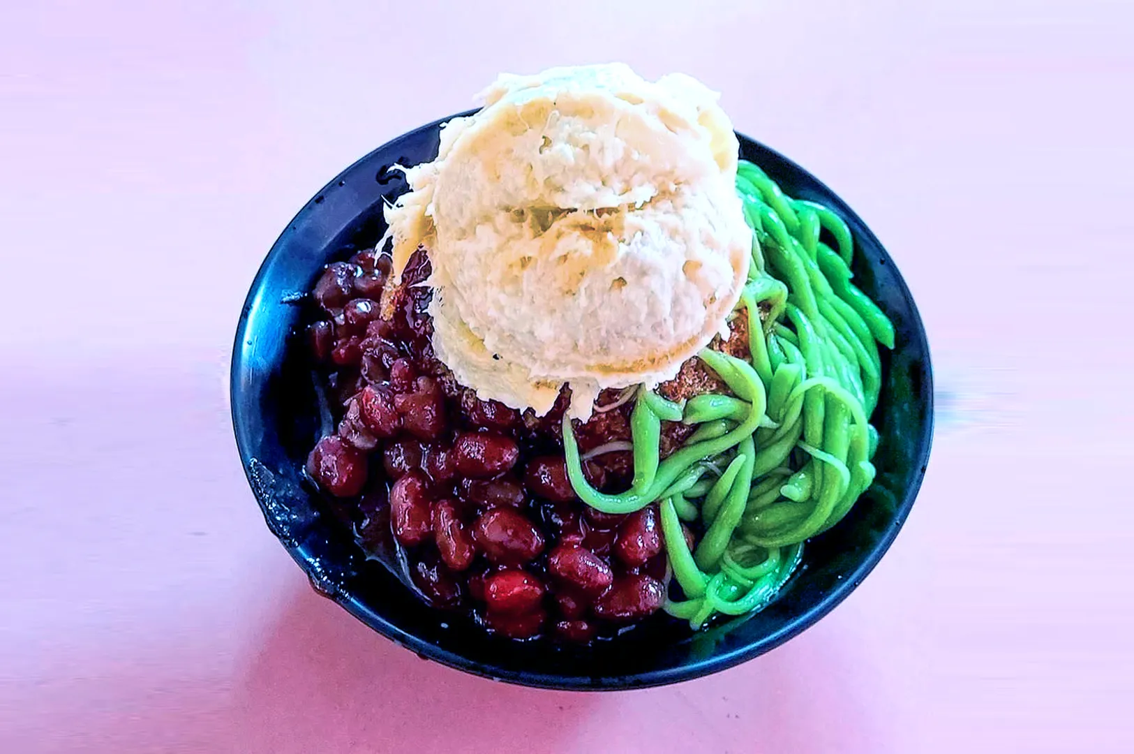A bowl of cendol with vibrant green jelly noodles, red beans, and a scoop of ice cream on top. The dish is colorful and looks refreshing.