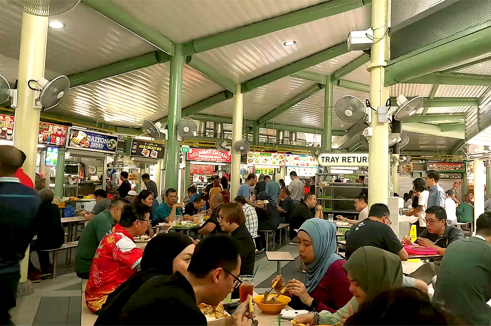 A bustling food court with diverse patrons seated at tables, enjoying meals. Food stalls with colorful signs are visible in the background, creating a lively atmosphere.