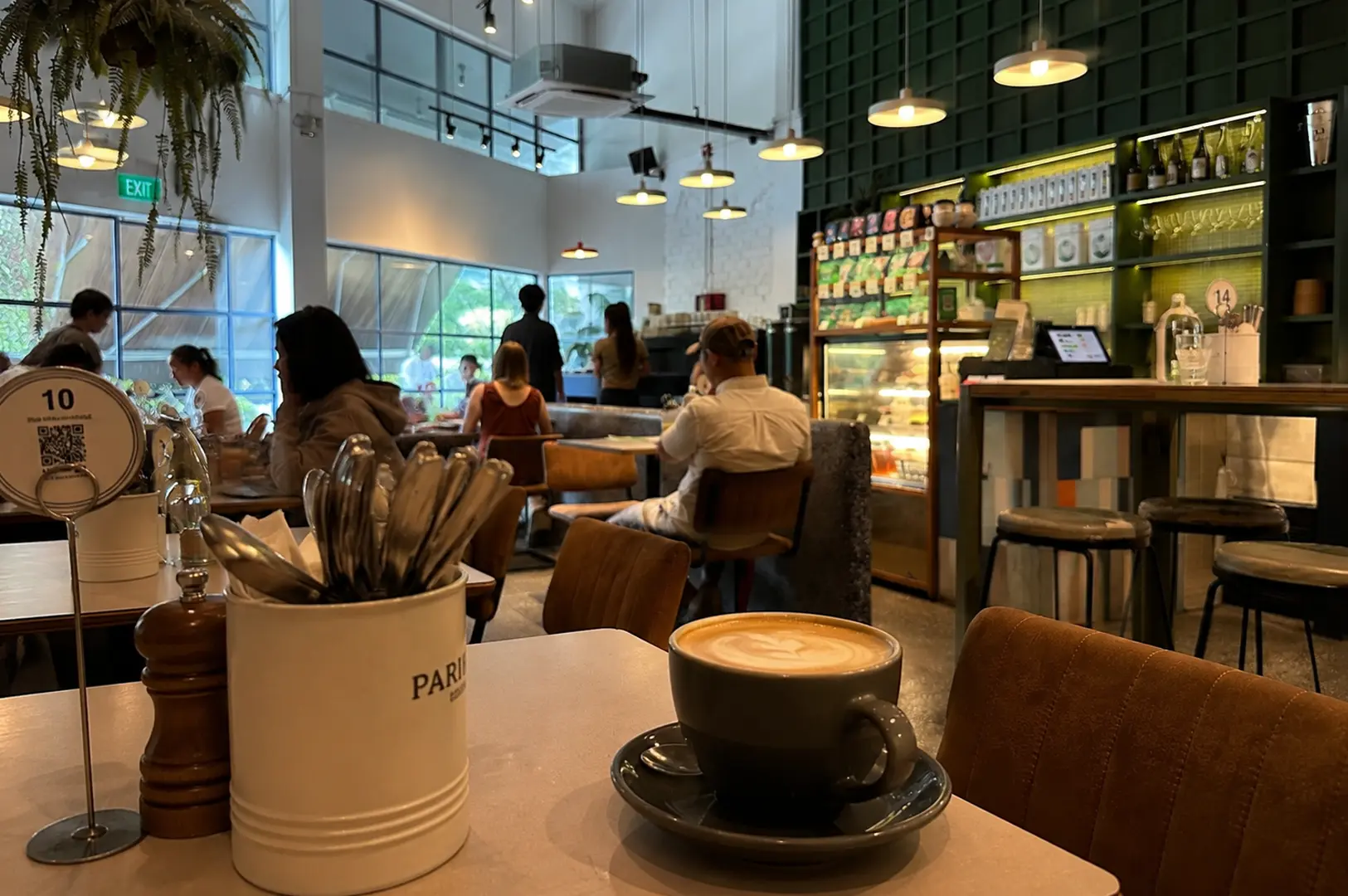 Latte coffee cup on table in foreground with cafe interior behind showing customers dining, green tiled walls, retail product shelves, table number stand, and hanging pendant lights