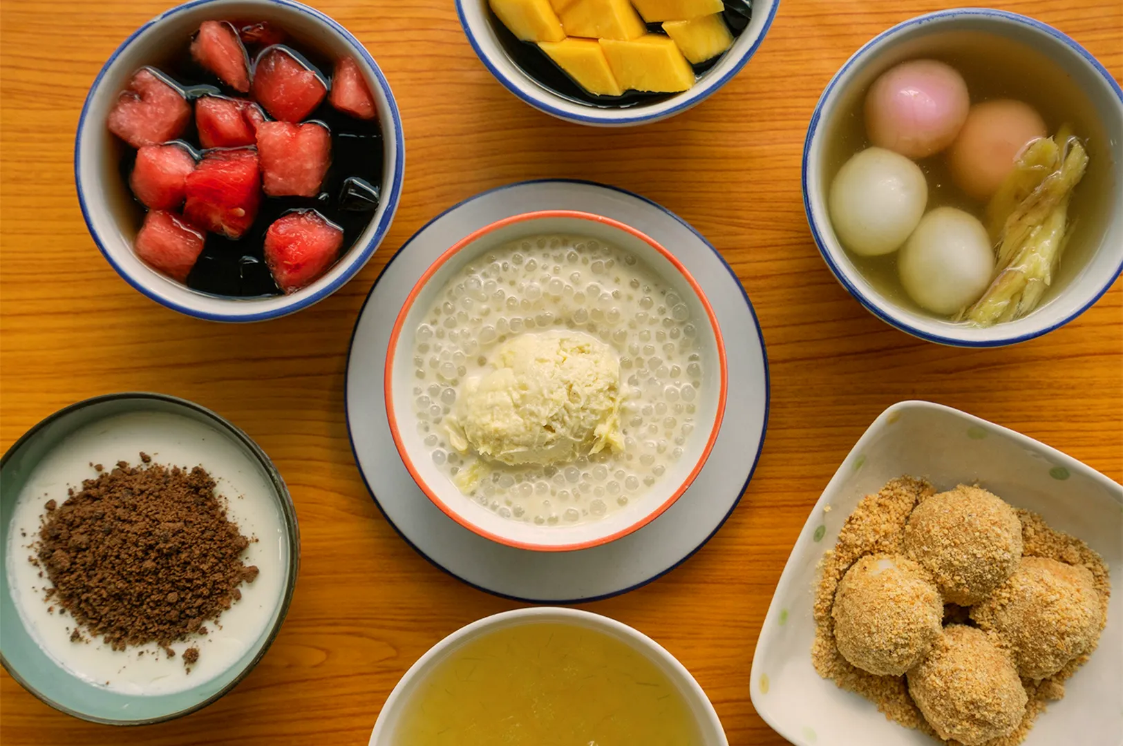 An assortment of colorful Asian desserts on a wooden table includes fruit bowls, sweet rice dumplings, shaved ice, and tapioca pudding, conveying a festive mood.