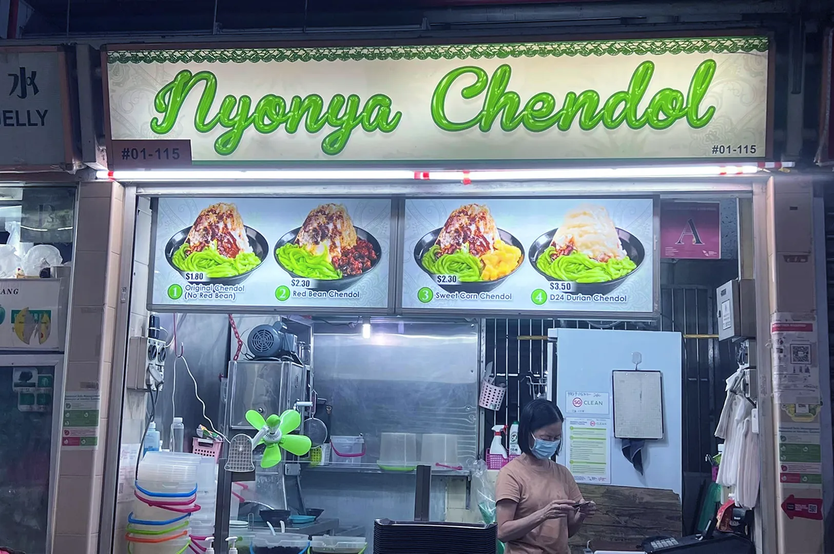Stall named "Nyonya Chendol" with a lit sign and three dessert images above, showing varieties of chendol. A person in a mask stands at the counter.