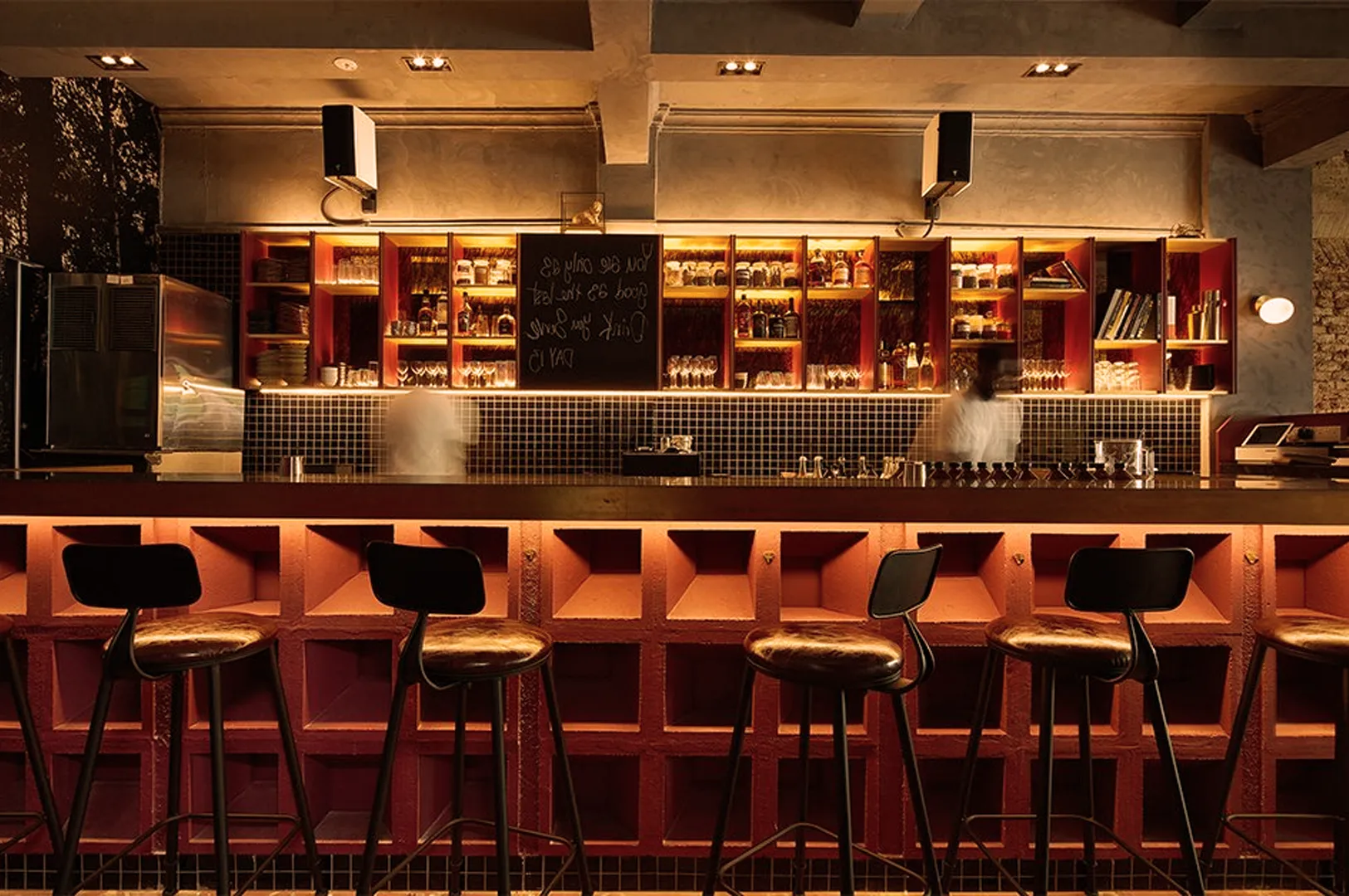 A cozy, dimly lit bar features a wooden counter with high stools and shelves filled with bottles. Warm lighting creates an inviting atmosphere