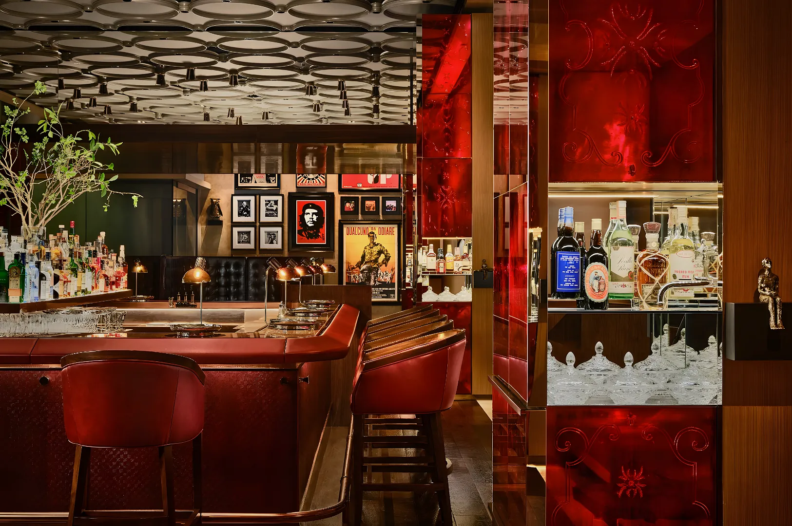 Stylish bar interior with a red leather counter, matching stools, and a variety of bottles on display. Framed artwork on walls adds a chic ambiance.