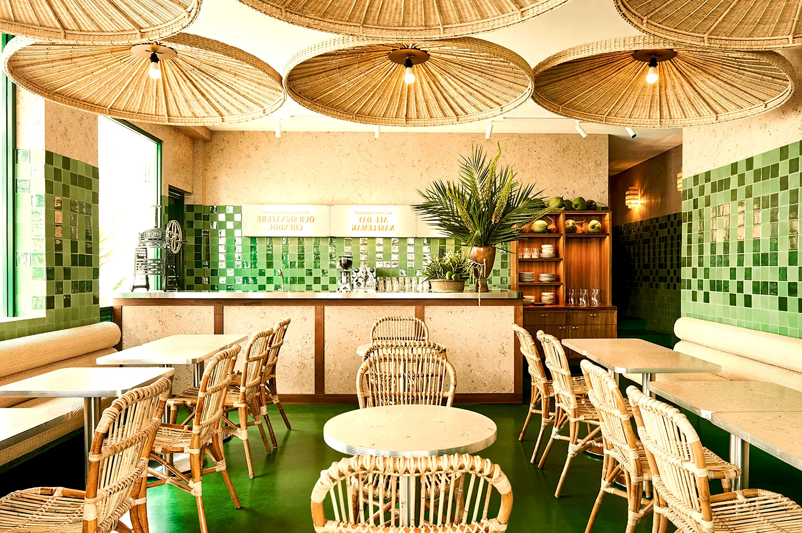 Cozy café interior with rattan chairs and round tables on a shiny green floor. Large woven pendant lights hang overhead, surrounded by green tiled walls.