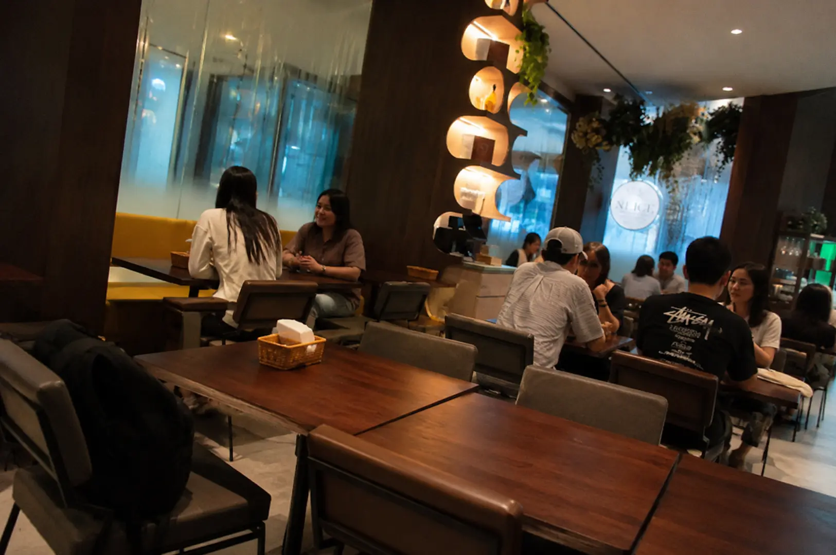 Cozy cafe interior with wooden tables and chairs, customers dining and conversing, decorative illuminated wall feature with circular cutouts and plants, large windows with blue-tinted glass