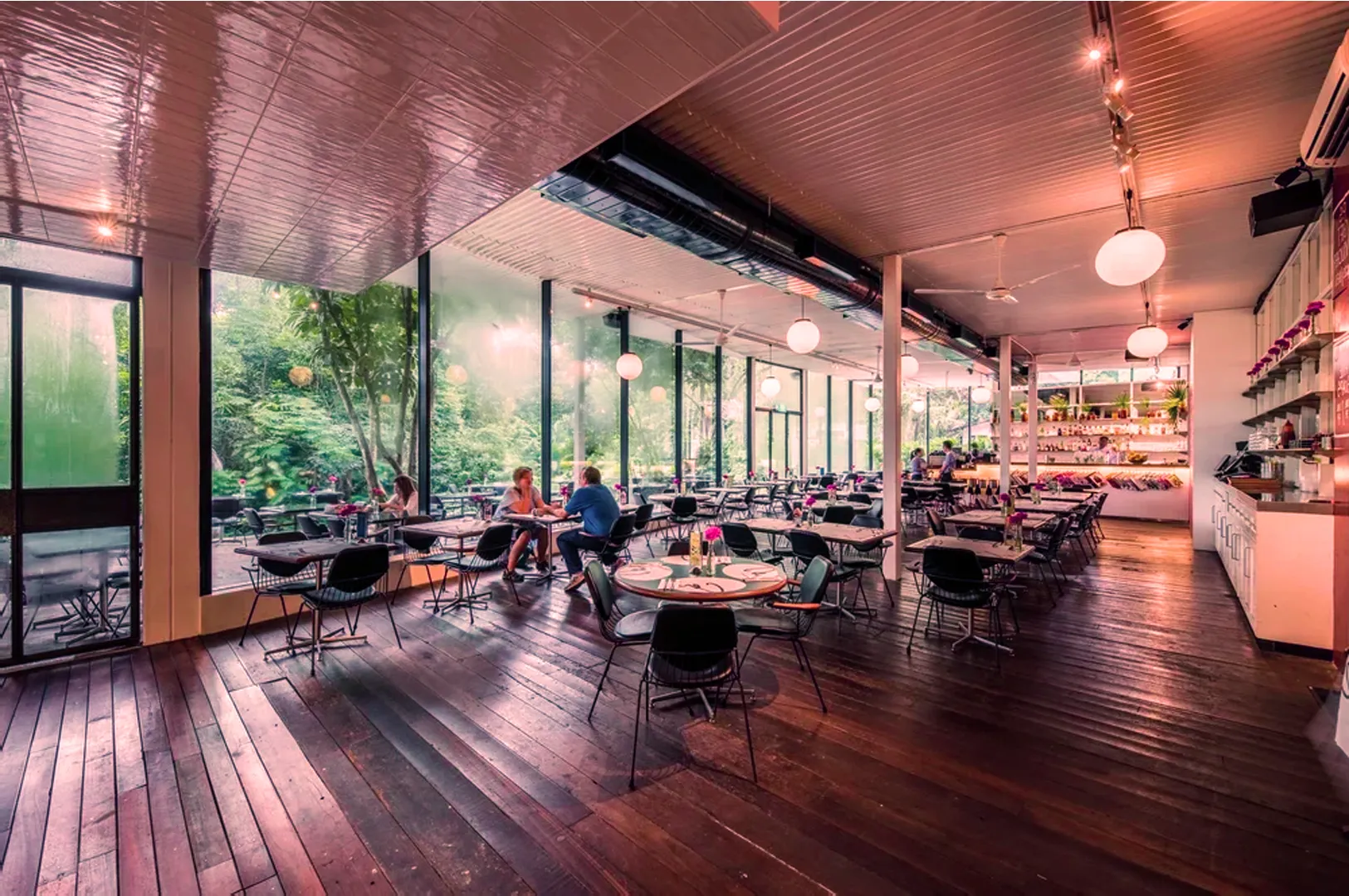 Spacious restaurant interior with wooden floors and large windows revealing greenery outside. Few people seated, warm lighting, and a relaxed ambiance.